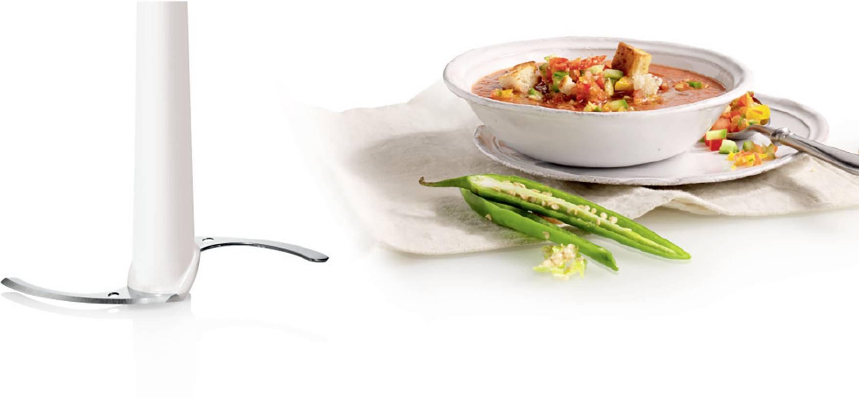 Bílý tyčový mixér vedle talíře se zeleninovou polévkou, ozdobené krutony a čerstvými zelenými chilli papričkami.