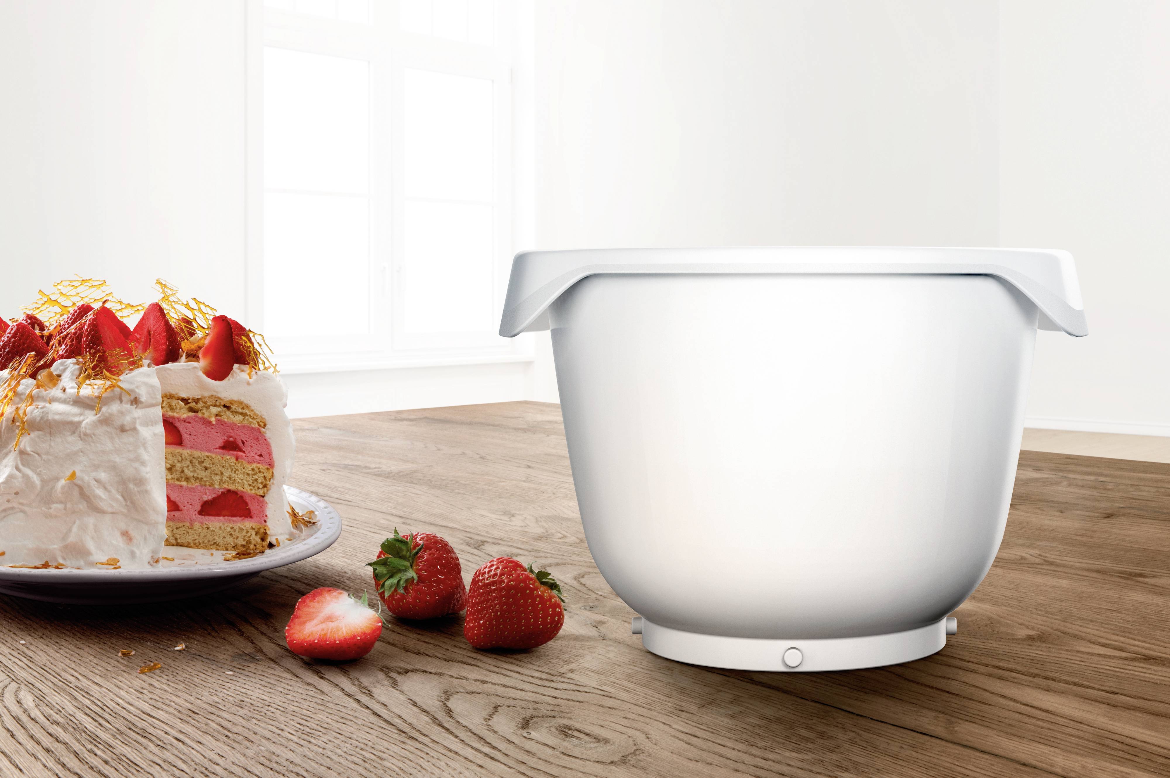 Bílá míchací miska stojí na dřevěném stole vedle talíře s jahodovým dortem. V pozadí je světlé okno.