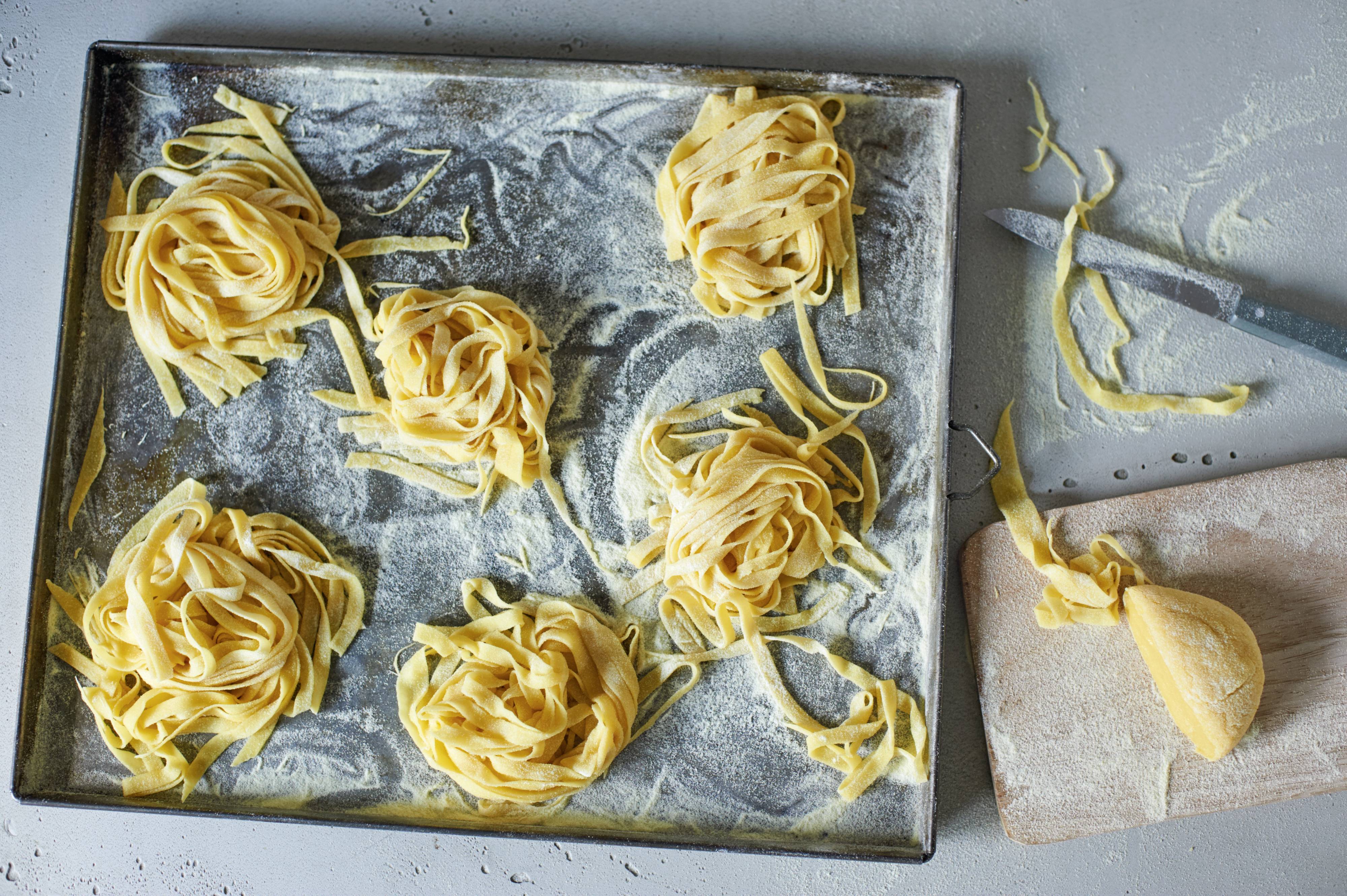 Domácí těstovinová hnízda a několik volných nudlí na moukou posypaném plechu. Vedle řezací nůž a kus těsta.