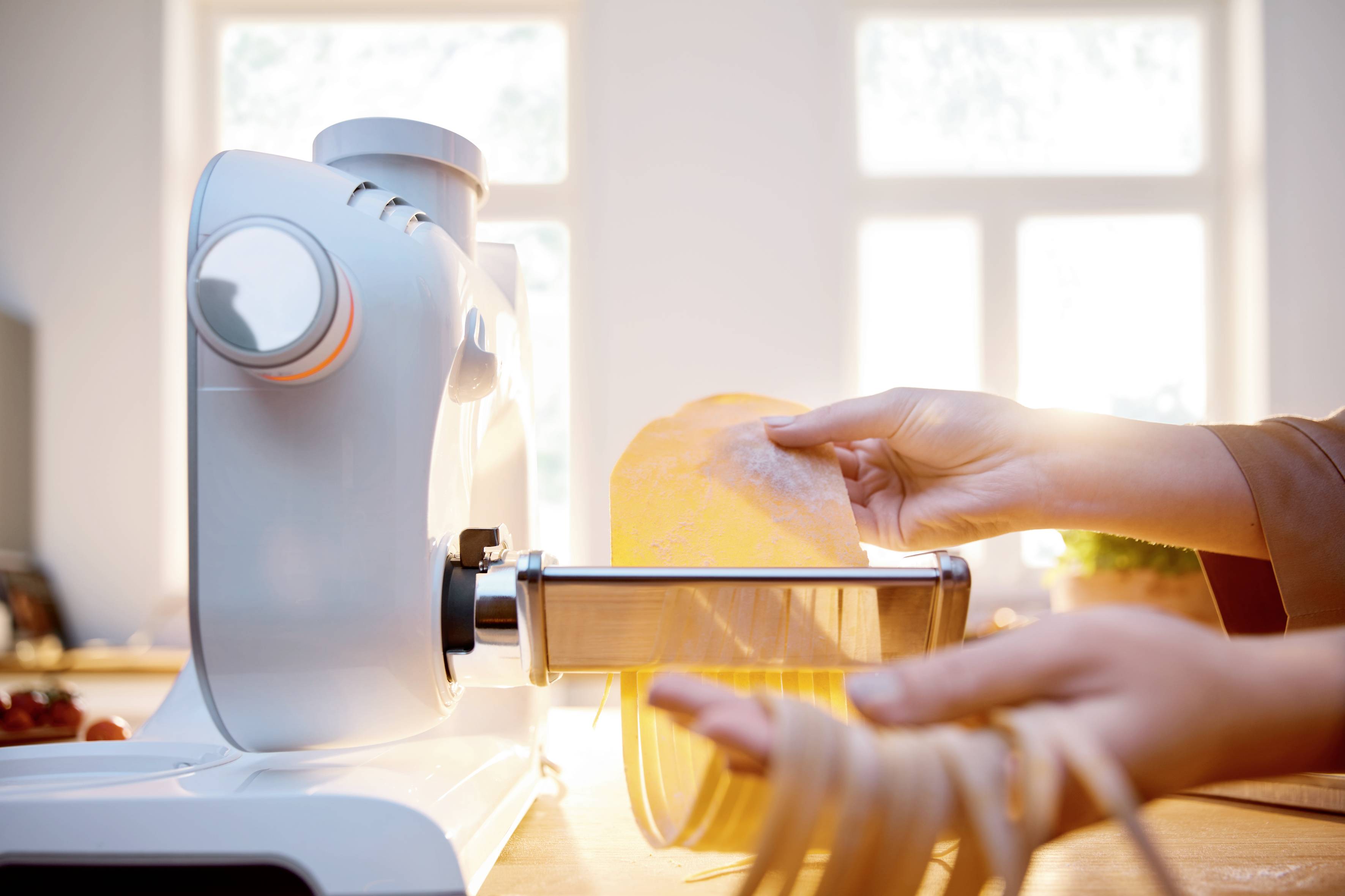 Jedna osoba používá strojek na těstoviny, aby vyrobila čerstvé těstoviny. Scéna je jasně osvětlena slunečním světlem procházejícím okny.