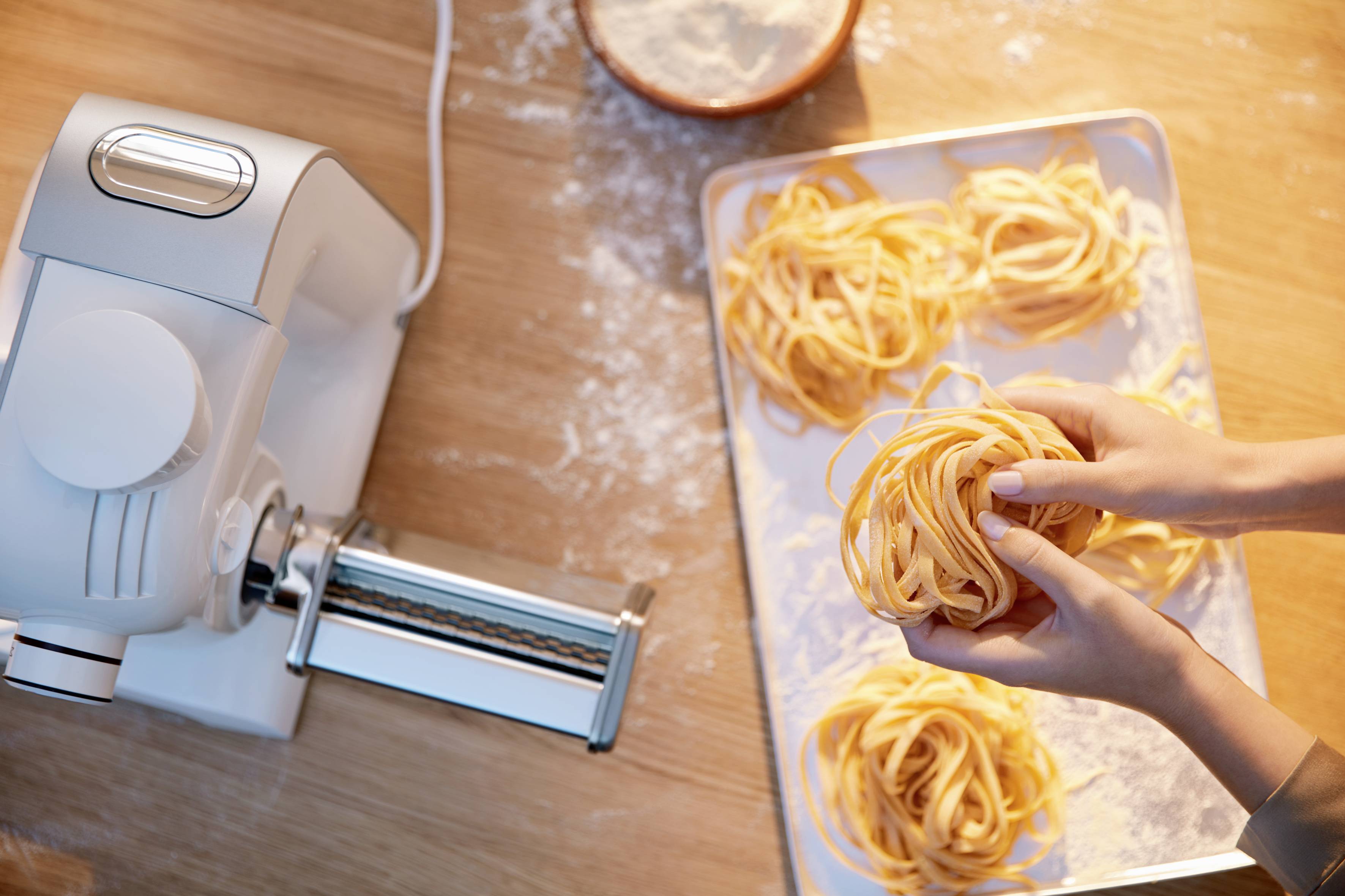 Osoba připravuje čerstvé těstoviny pomocí strojku na těstoviny a mouky na dřevěném stole. Ruce vytváří hnízdo z těstovin.