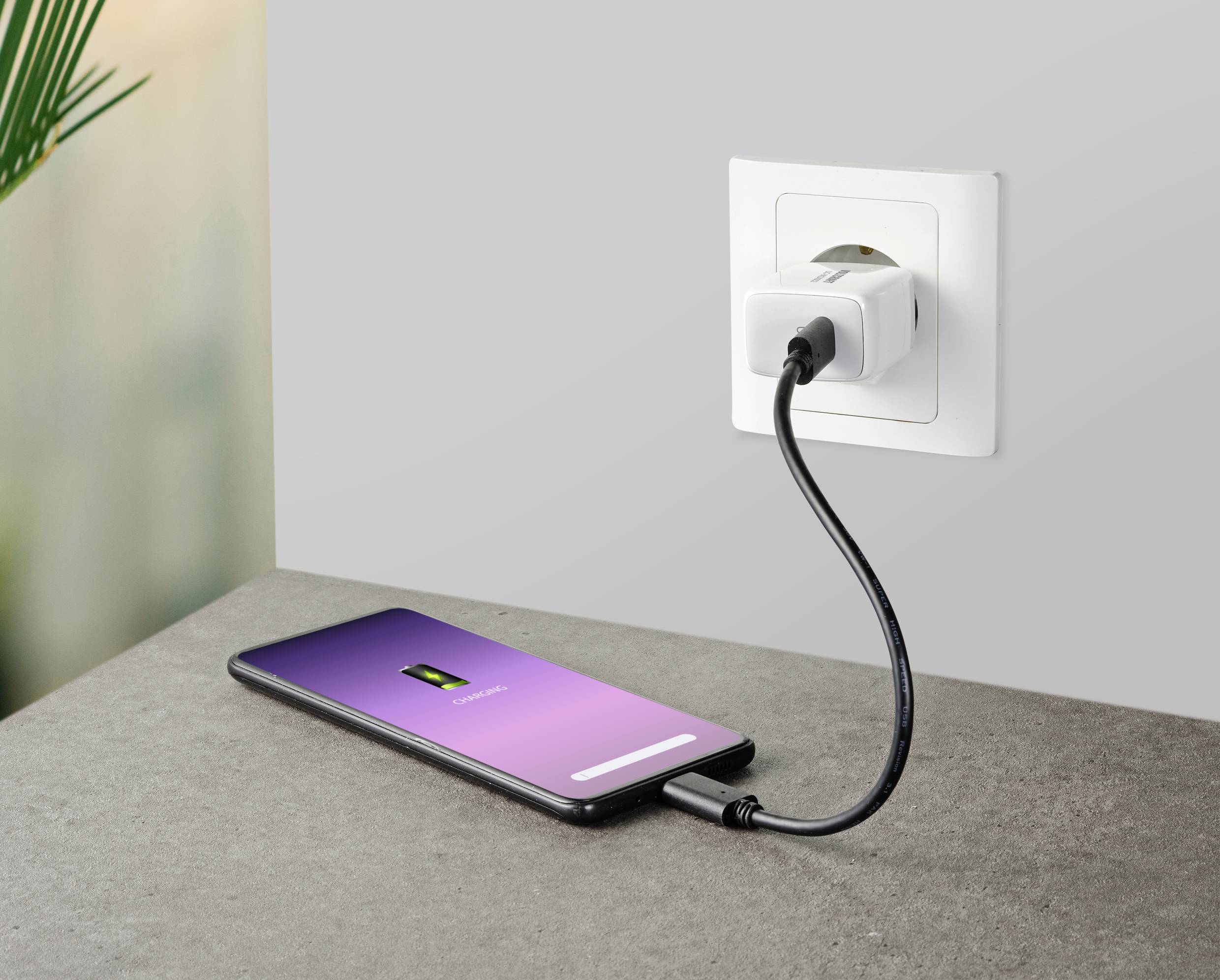 Smartphone na stole, připojené k elektrické zásuvce nabíjecím kabelem. Na displeji je zobrazen symbol baterie a probíhá nabíjení.