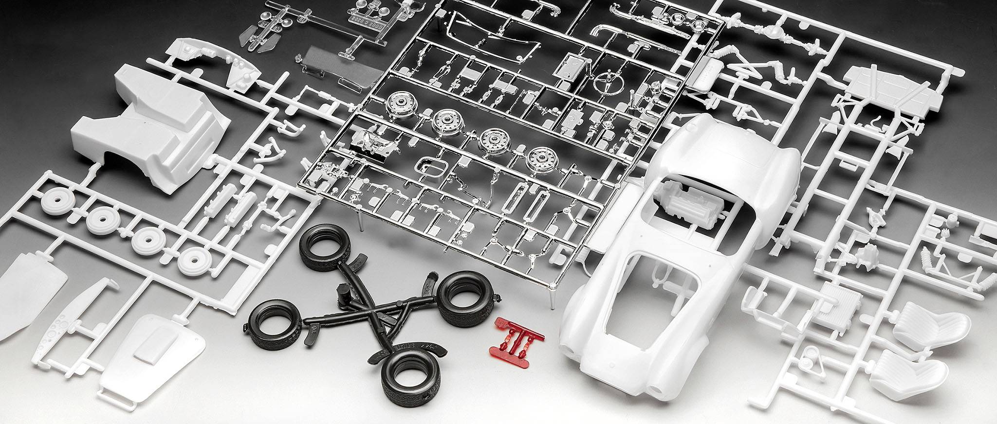 Plastikový modelářský kit s díly pro automobil: karoserie, kola a další komponenty rozmístěné na šedém pozadí.