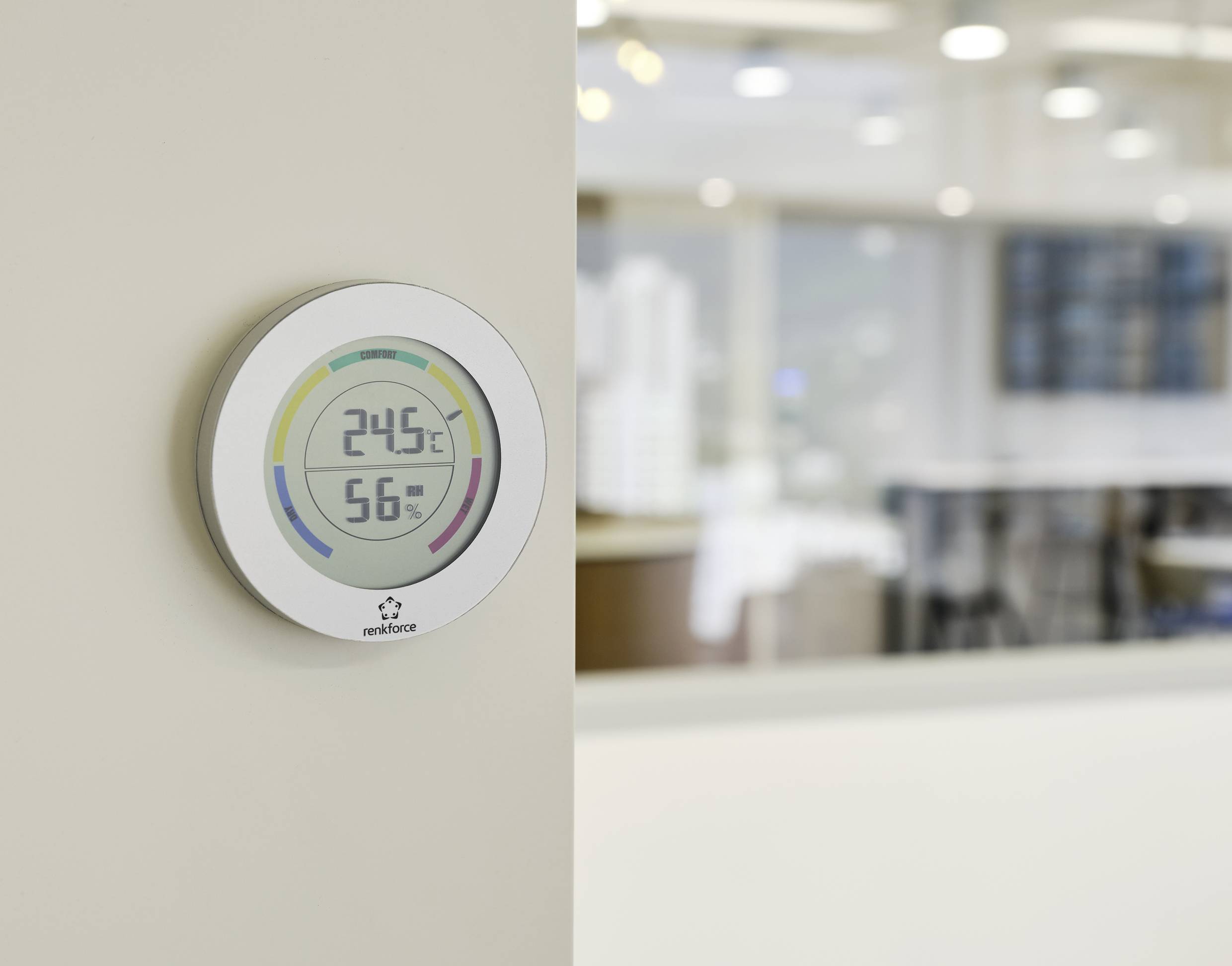 Digitální termostat na stěně ukazuje teplotu 24,5 °C a vlhkost vzduchu 56 %, v pozadí je kancelář.