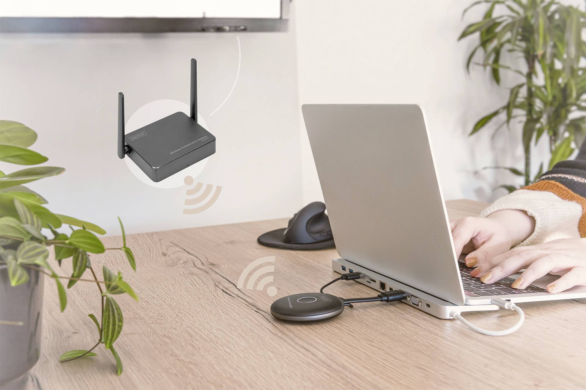 Osoba píše na notebooku připojeném k bezdrátovému adaptéru; v pozadí router vysílající bezdrátové signály.