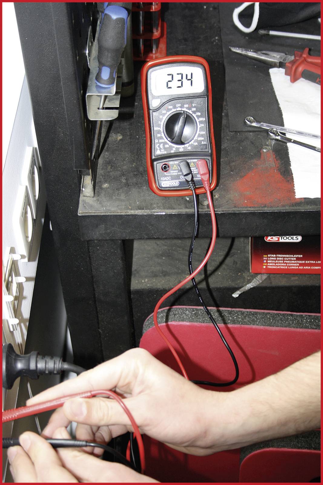 Multimetr měří elektrické napětí 234 voltů. V popředí drží ruka měřicí kabely připojené k přípojce. Prostředí: dílna.