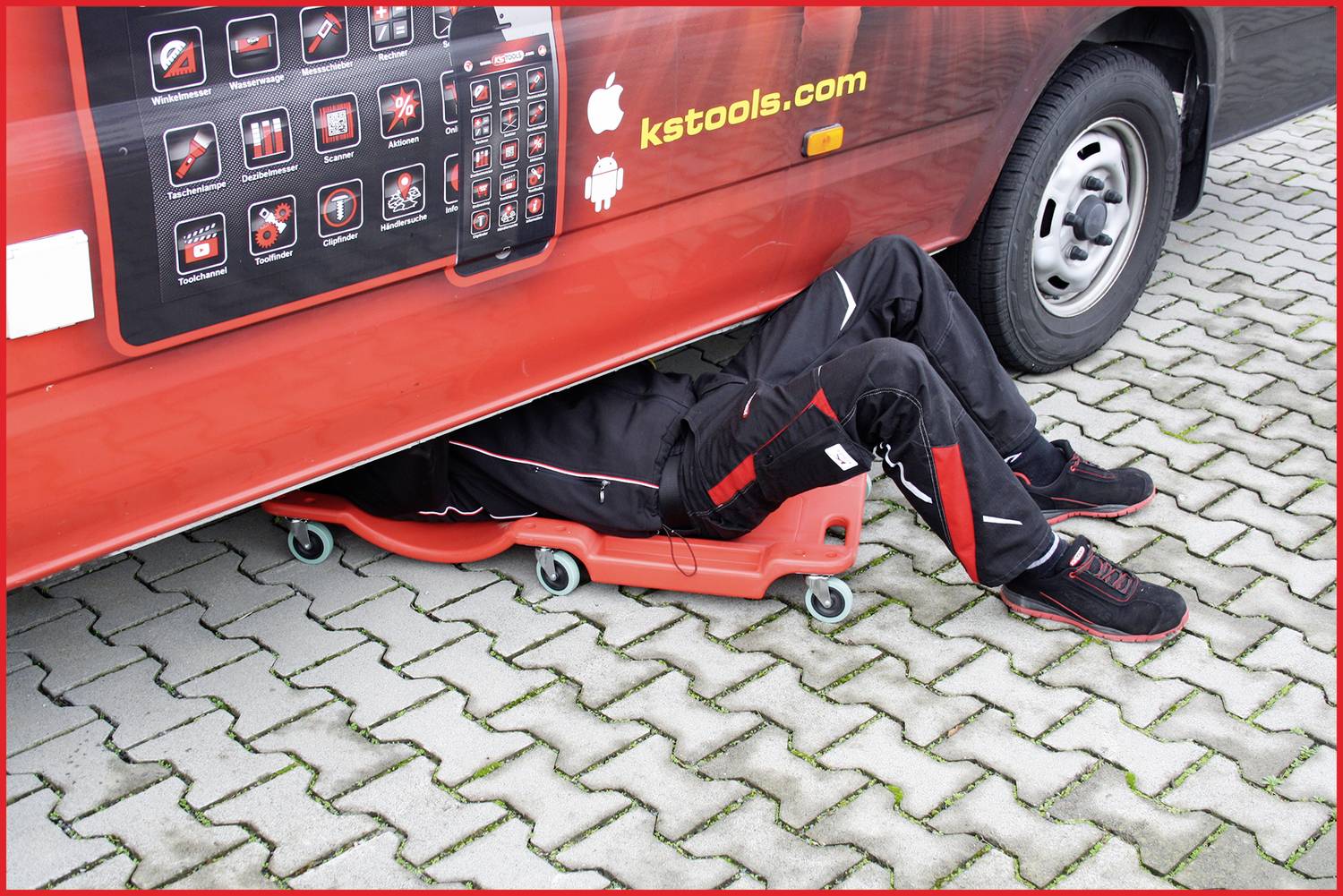 Mechanik leží na montážním lehátku pod červeným dodávkovým vozem na dlážděném parkovišti.