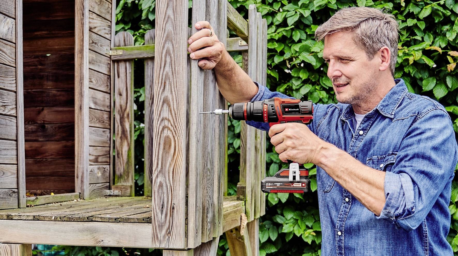 Muž vrtá dřevěné latě pomocí elektrické vrtačky. V pozadí je vidět dřevěná konstrukce.