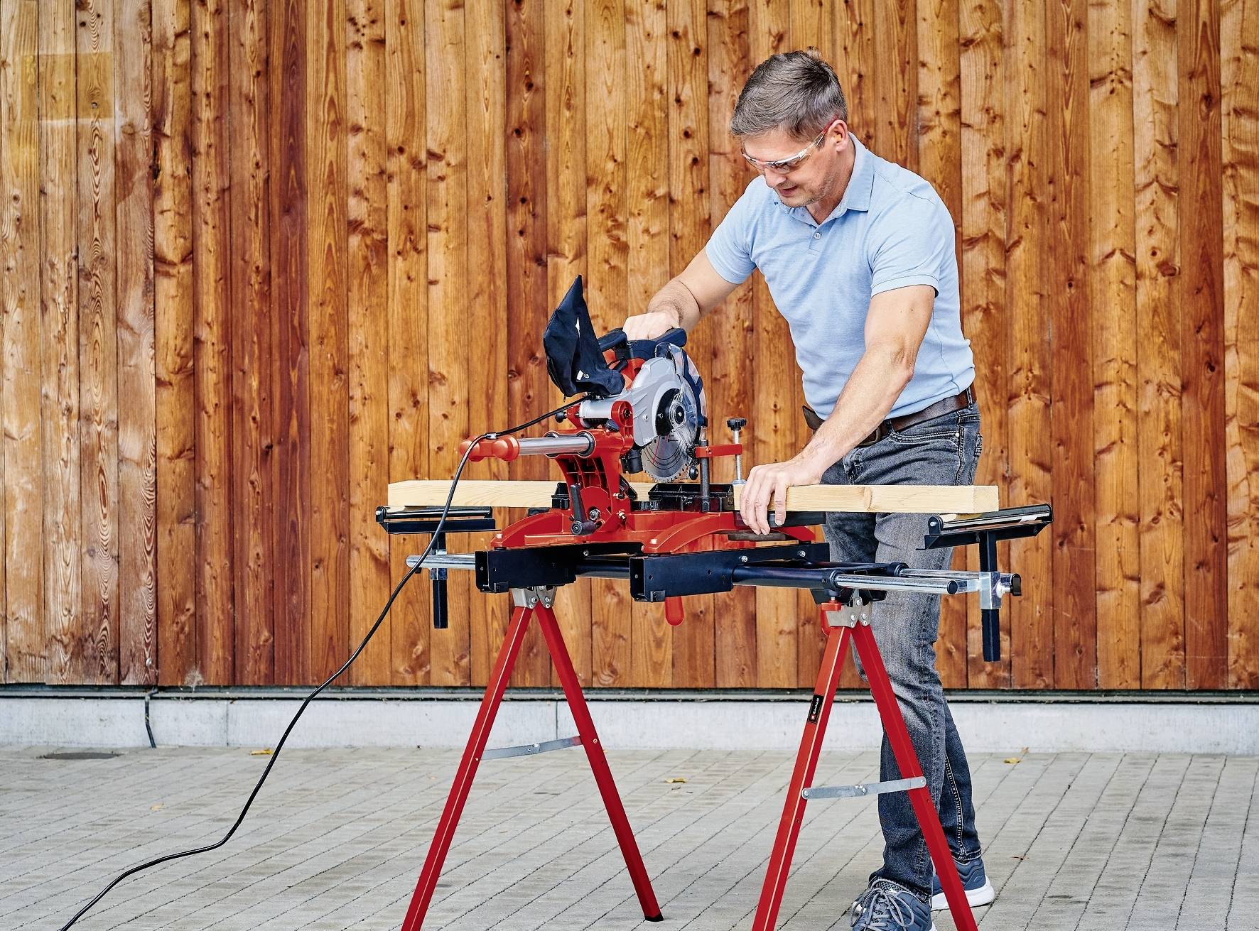 Muž v ochranných brýlích používá stolní kotoučovou pilu k řezání dřevěné desky. V pozadí je vidět dřevěná stěna.