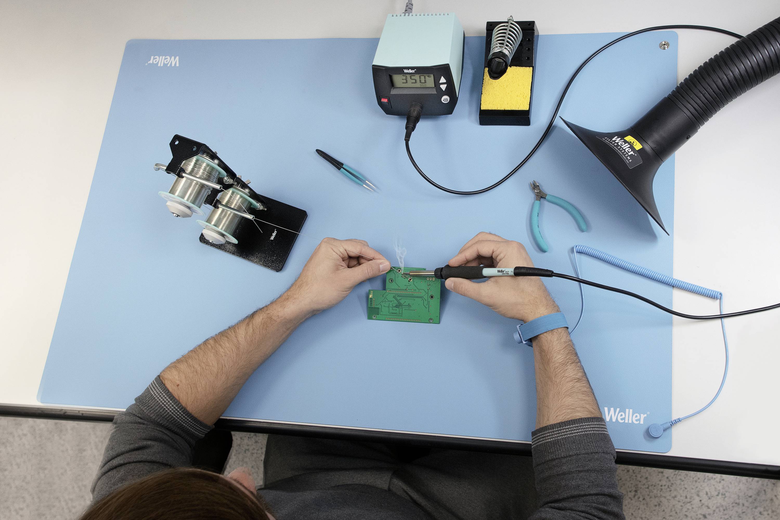 Osoba pájí elektronické komponenty na zelené desce plošných spojů u pracovního stolu, obklopena nástroji a vybavením.