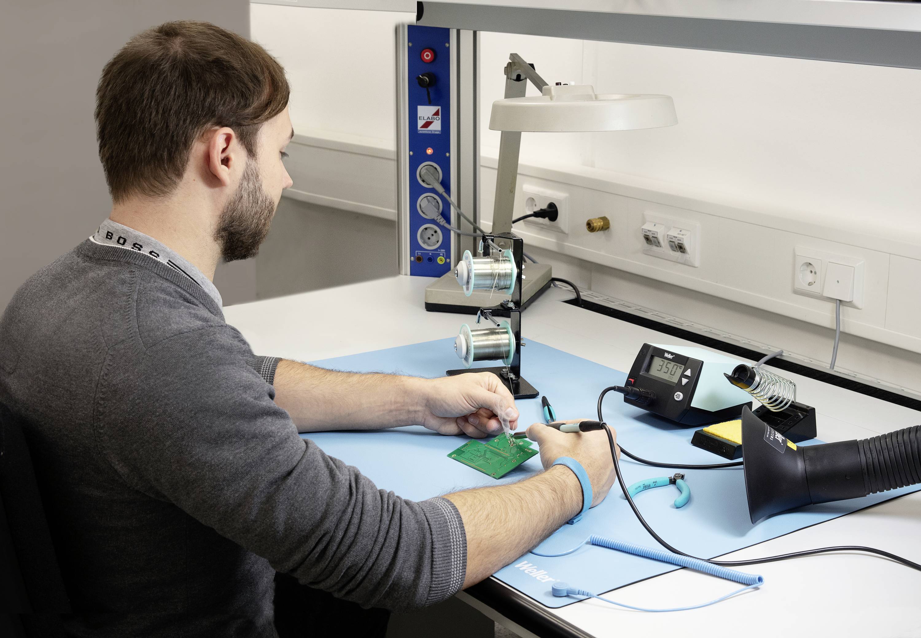 Osoba pájí zelenou desku plošných spojů u pracovního stolu s elektronickým vybavením a nástroji.