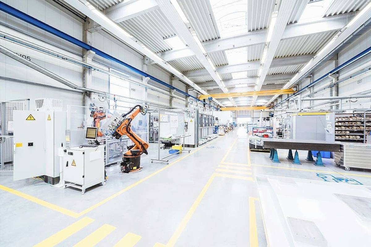 Průmyslová hala s automatizovaným robotickým ramenem, osvětlená horními světlíky. Prostředí obklopené stroji a hotovými výrobky.