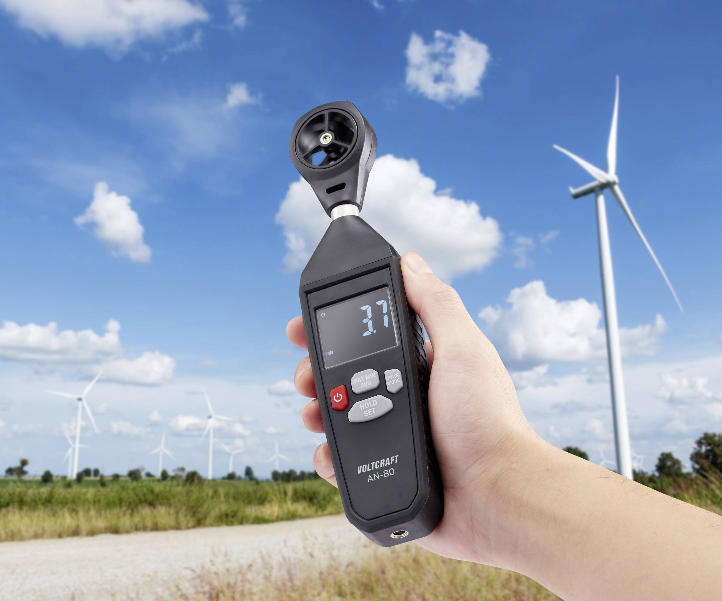 Jedna ruka drží anemometr, který ukazuje rychlost větru 3,7. V pozadí je vidět několik větrných turbín.