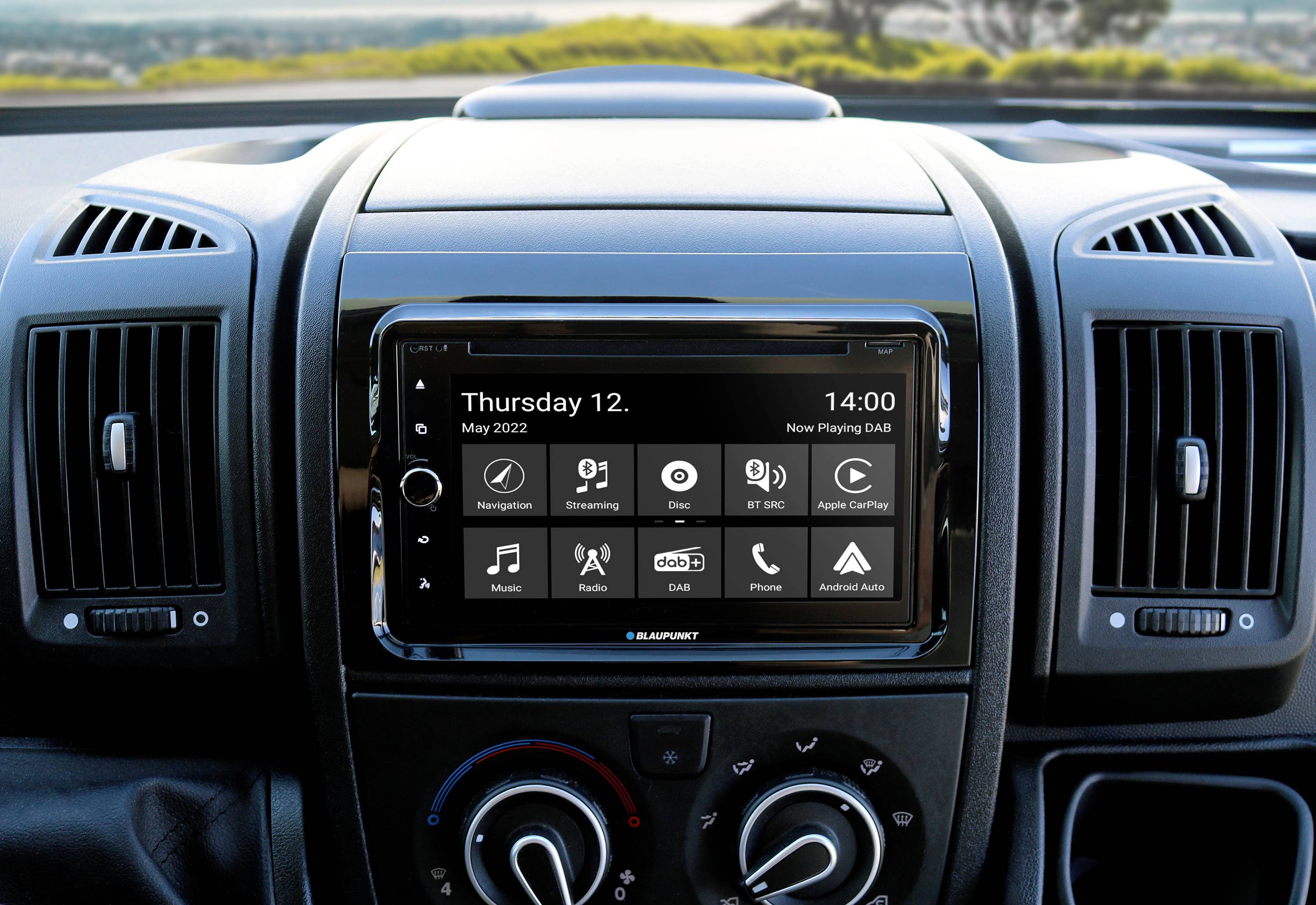Palubní displej automobilu s datem 'Čtvrtek, 12. května 2022', časem '14:00' a ikonami aplikací pro navigaci, rádio a hudbu.