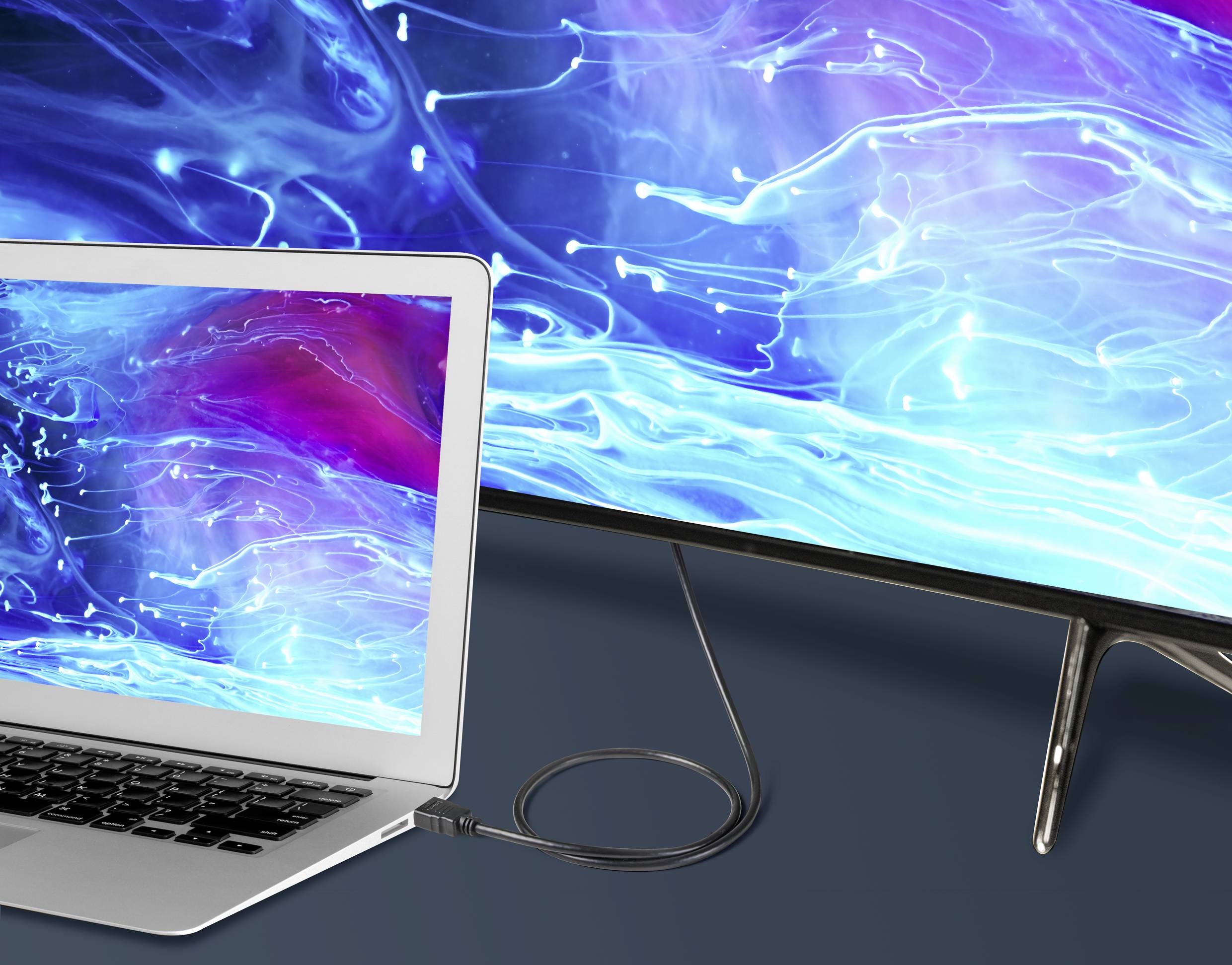 Notebook připojený kabelem k velkému monitoru. Oba displeje zobrazují abstraktní modrý vzor.