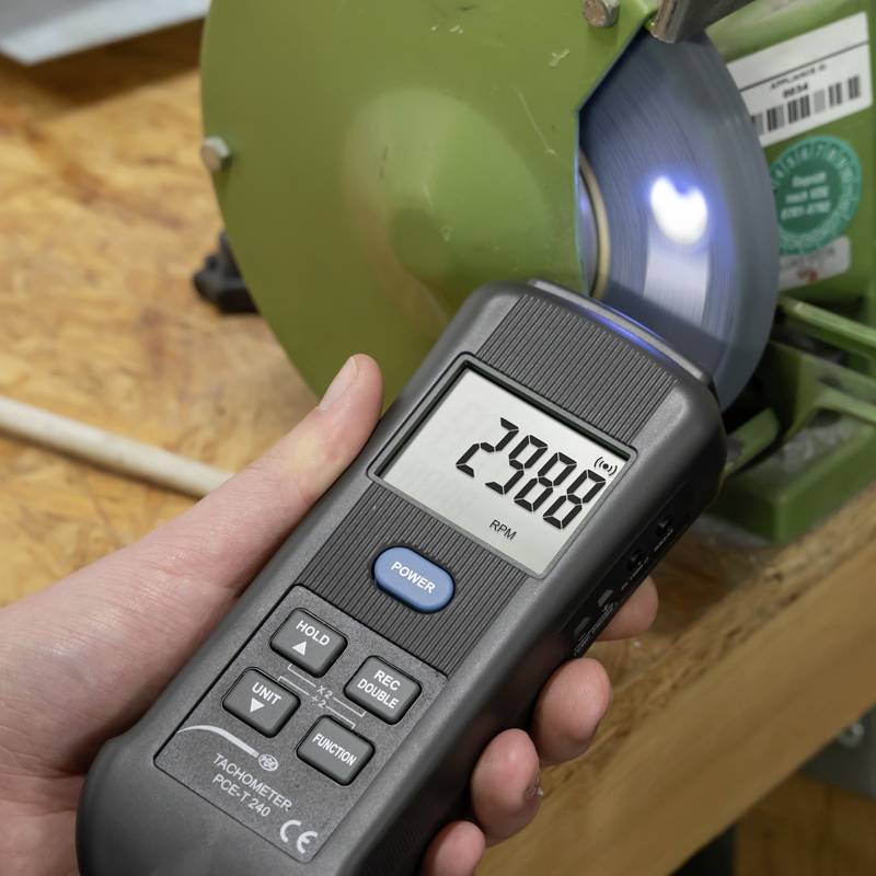 Ruka měří otáčky rotující brusné desky pomocí tachometru. Přístroj ukazuje '2988 ot./min'.