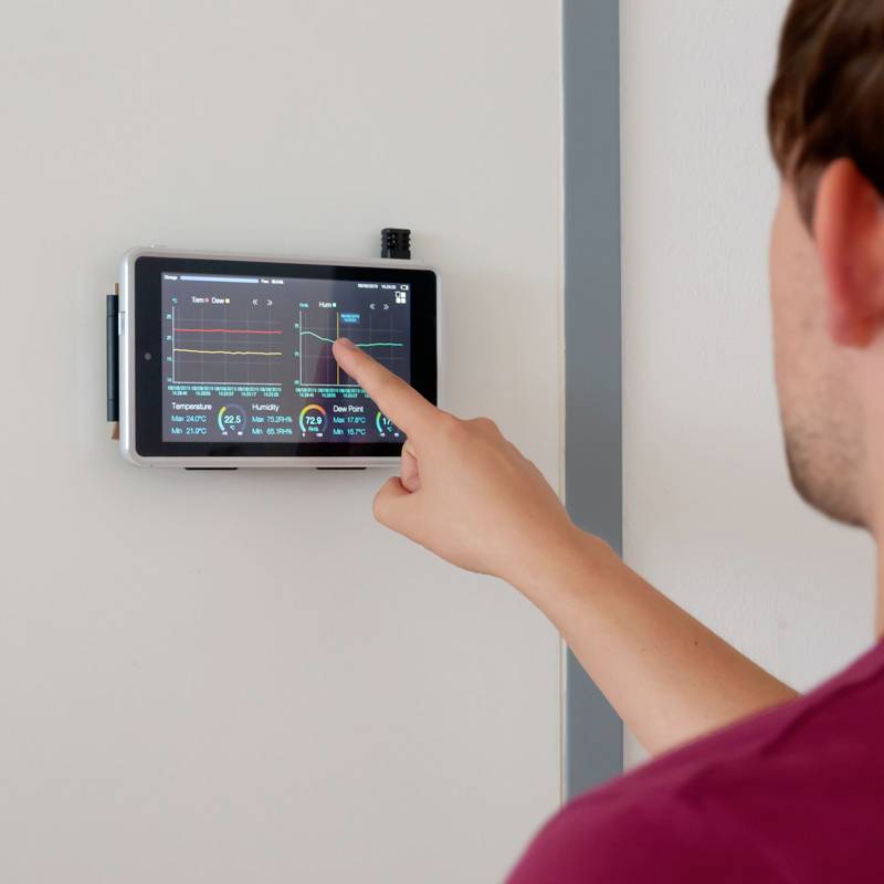 Osoba obsluhuje nástěnný tablet pro ovládání inteligentního domácího systému, který zobrazuje údaje o teplotě a spotřebě energie.