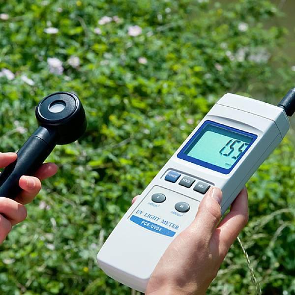 Dvě ruce drží digitální měřicí přístroj v zeleni, který ukazuje hodnotu UV indexu 0,55. Senzor je připojen kabelem.