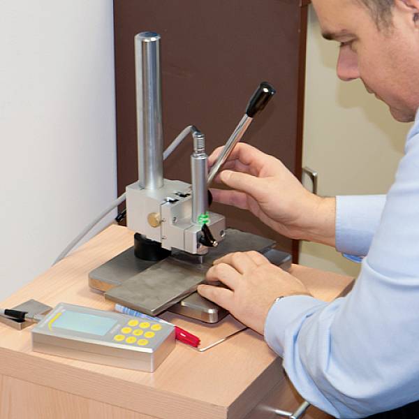 Muž obsluhuje kovové přesné zařízení na stole, které je pravděpodobně používáno v laboratoři nebo dílně.