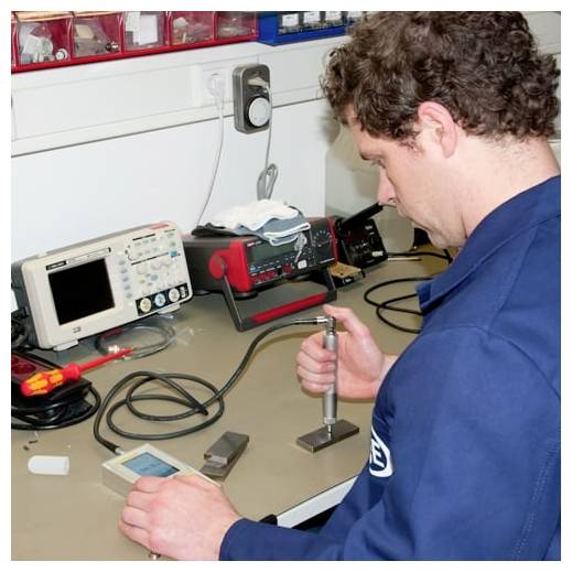Technik testuje elektronické zařízení na pracovní ploše, přičemž drží sondu podobný nástroj vedle laboratorního vybavení a kabelů.