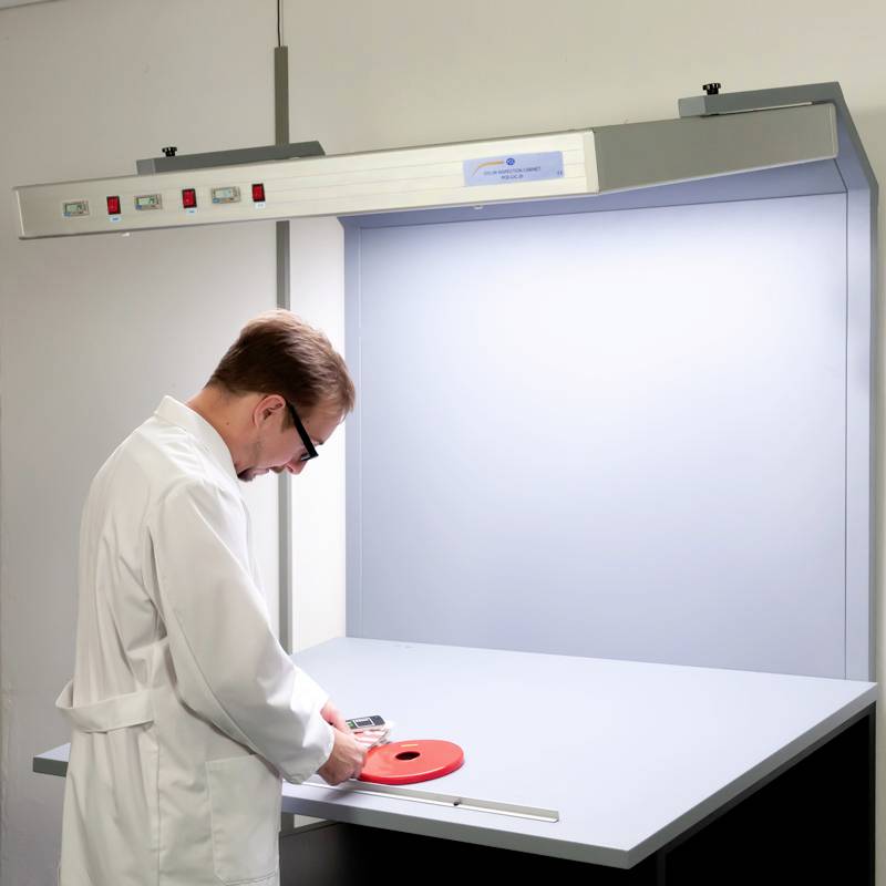Osoba v bílém laboratorním plášti pracuje u osvětleného stolu a zkoumá červený objekt. Pozadí je bílé, prostředí technické.