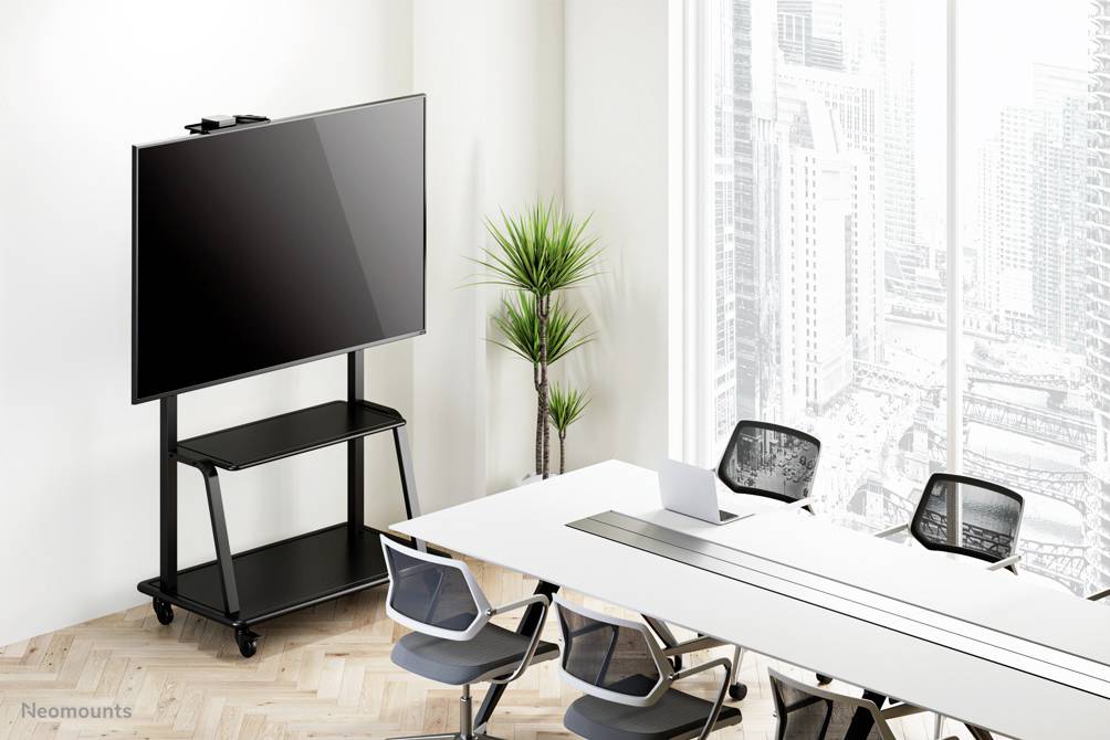 Moderní konferenční místnost s velkým displejem na mobilním stojanu, bílým stolem a čtyřmi židlemi před prosklenou stěnou.