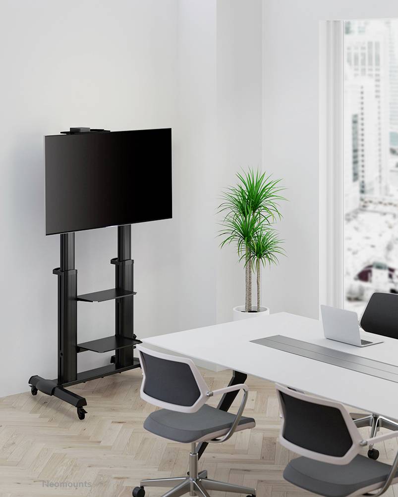 Moderní konferenční místnost s velkým monitorem na stojanu, bílým stolem a třemi kancelářskými židlemi. V pozadí stojí pokojová rostlina v květináči.