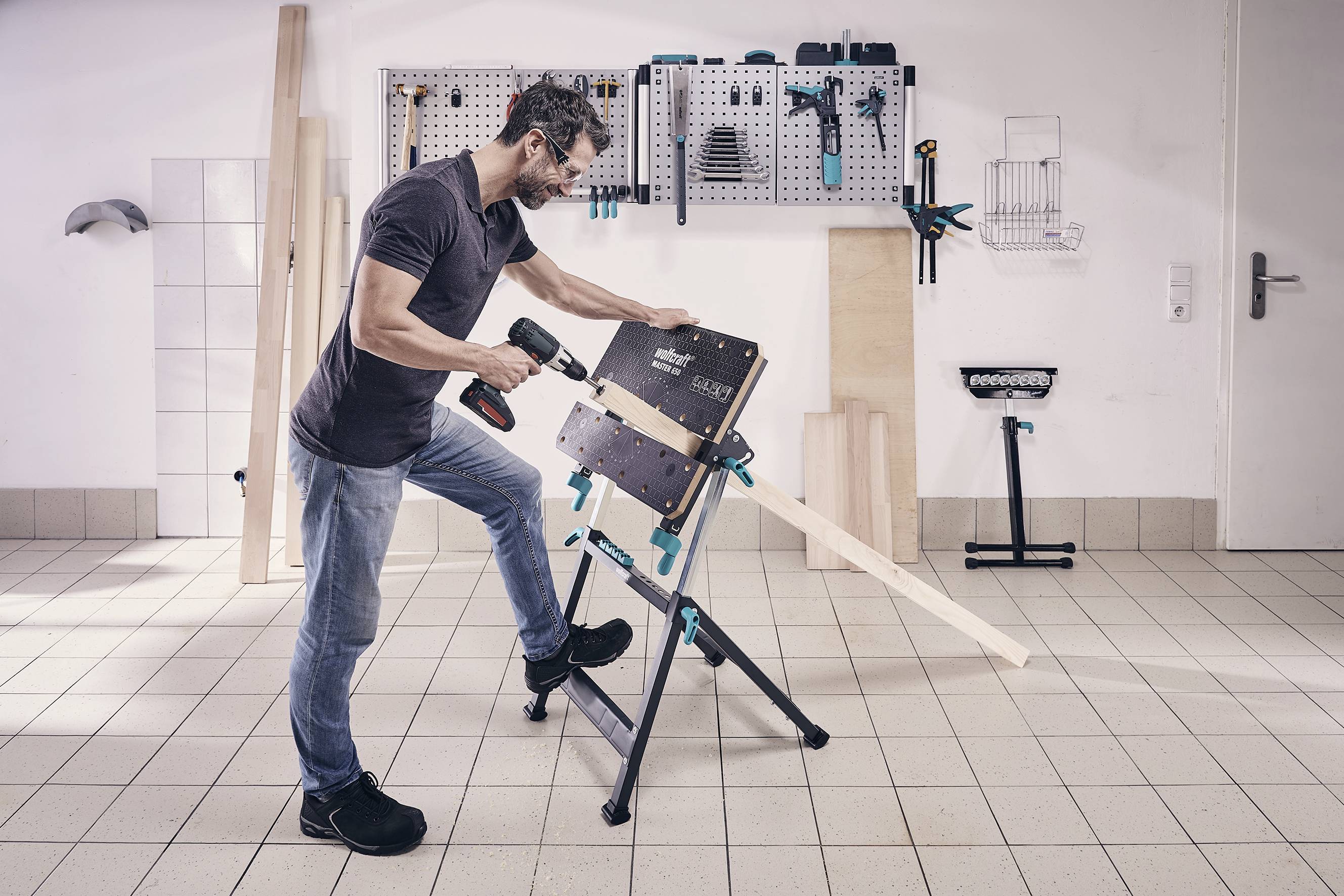 Muž pracuje v dílně na kusu dřeva, který je upevněný ve svěráku. Používá elektrickou vrtačku. V pozadí jsou nástroje zavěšené na stěně.