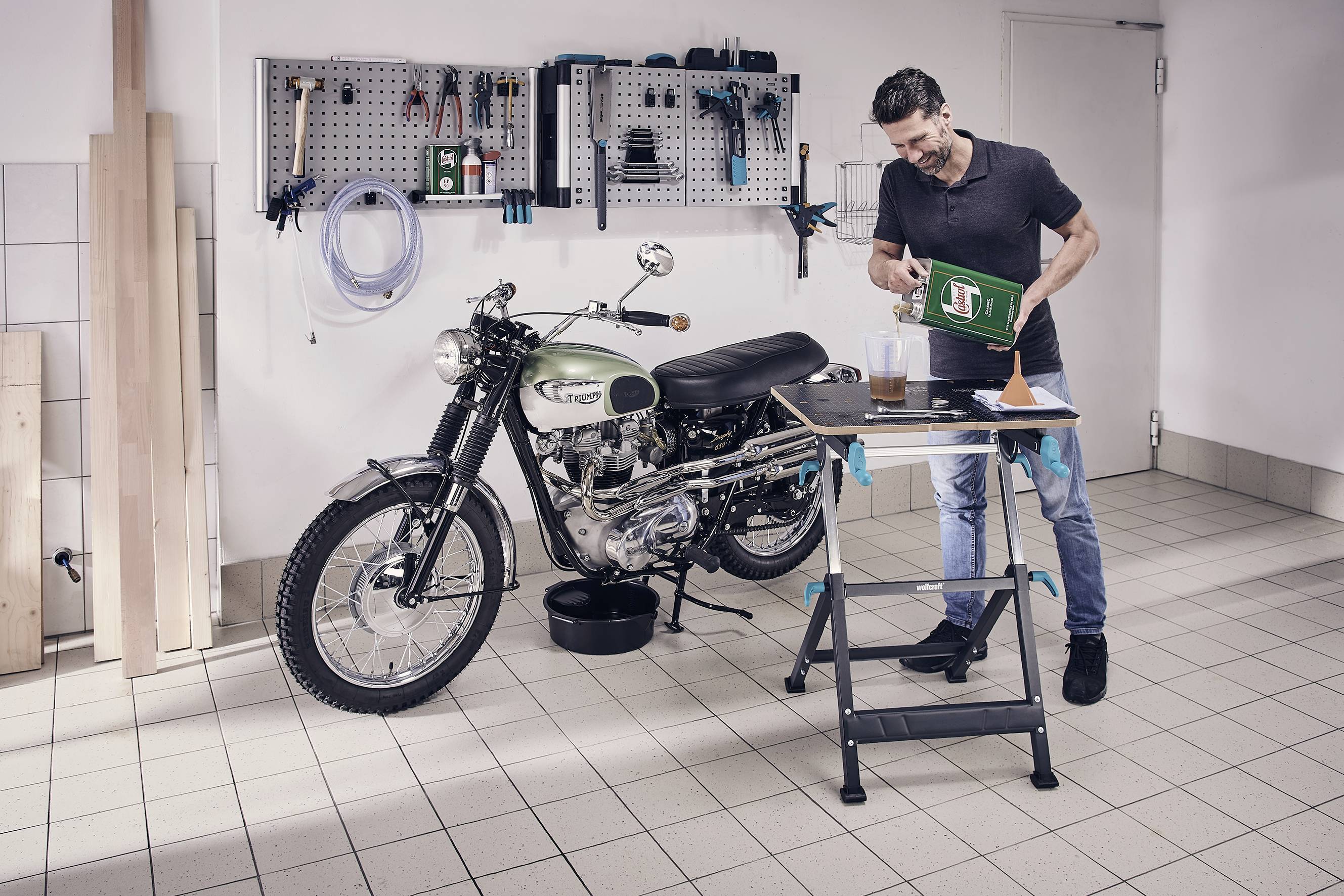 Muž v dílně nalévá olej do motocyklu. Nářadí a příslušenství na stěně. Zaměření na údržbu motocyklu.