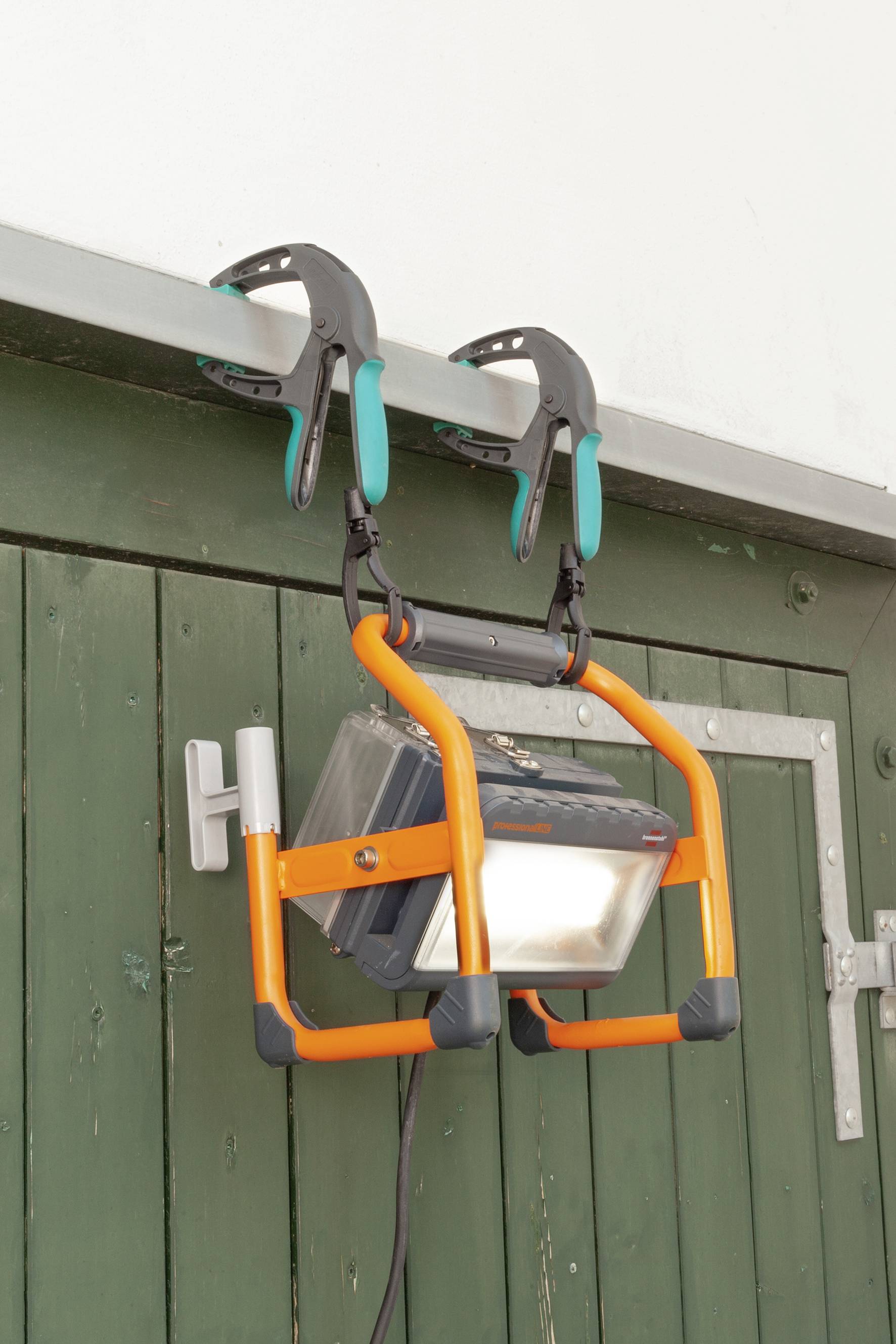 Přenosná pracovní lampa je připevněna dvěma svorkami na zelených dřevěných dveřích. Má oranžový rámeček.