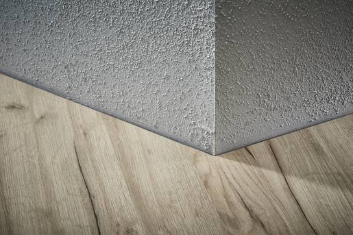 Roh místnosti s strukturovanou, šedou omítkou na stěně a dřevěnou podlahou. Textury jsou zřetelně viditelné.