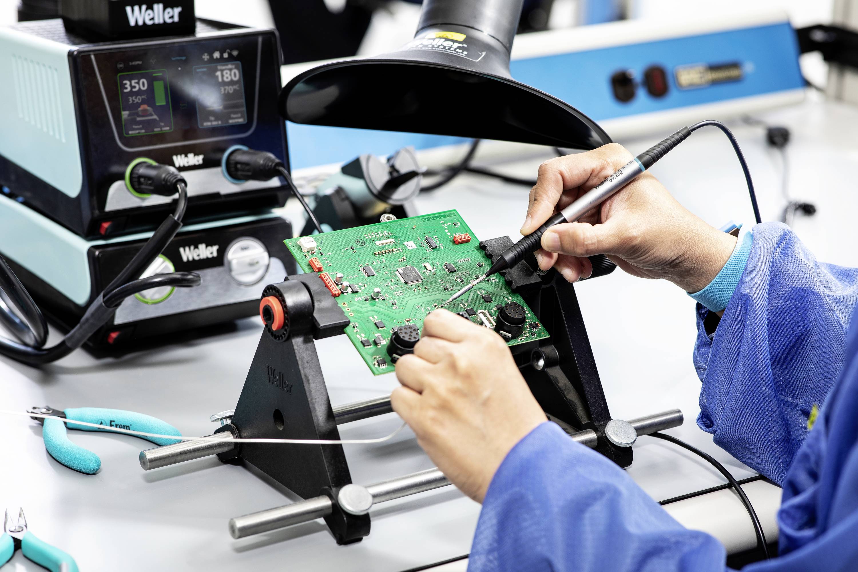 Osoba pájí na zelené desce plošných spojů pomocí nástrojů. V pozadí pájecí stanice s digitálním displejem.