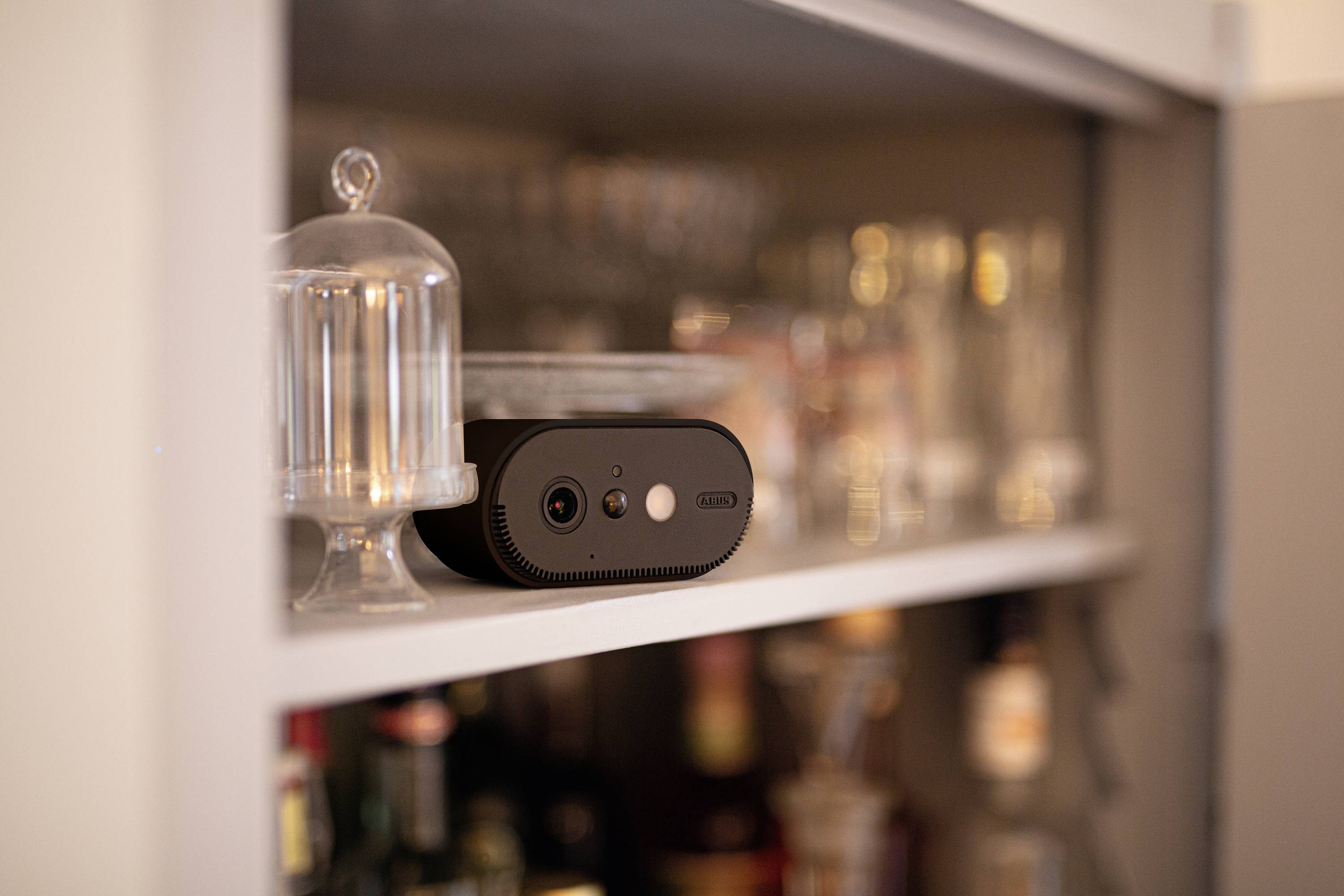 Malá černá kamera stojí na polici v kuchyni, obklopená sklenicemi. Slouží k monitorování nebo nahrávání.