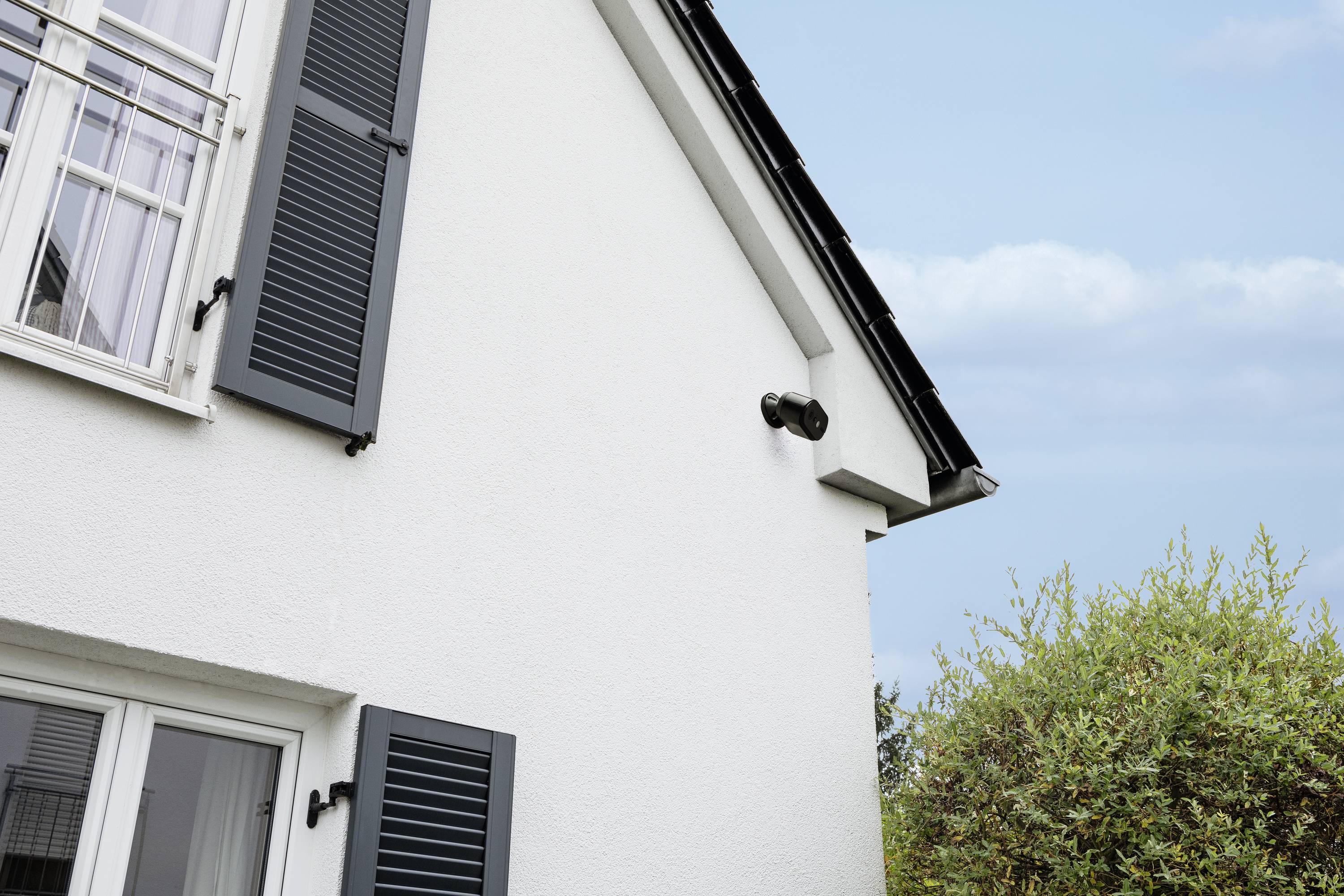 Bílá budova s modrými okenicemi, bezpečnostní kamera na vnější straně, vedle ní strom a modrá obloha v pozadí.