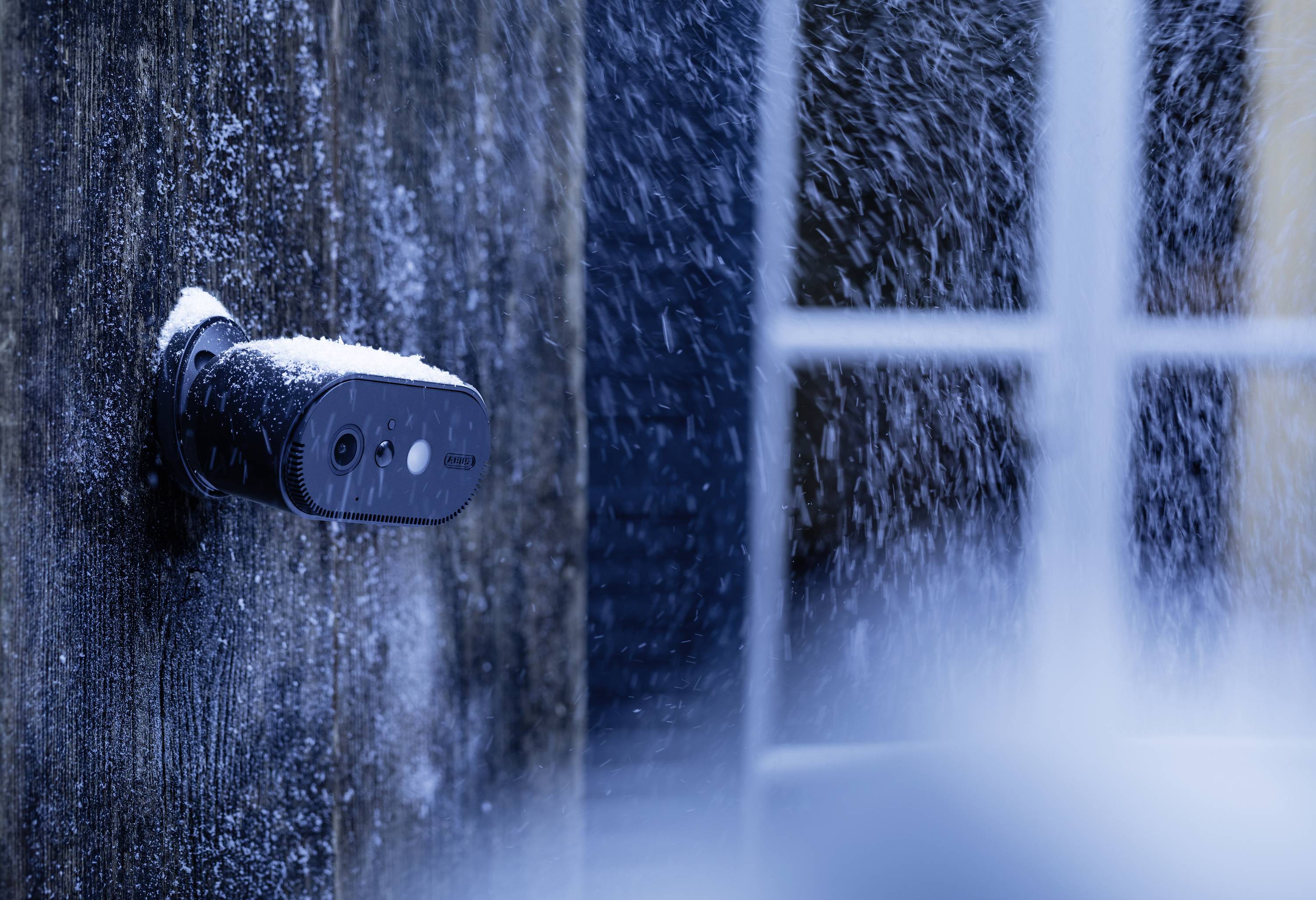 Bezpečnostní kamera na dřevěné stěně, pokrytá sněhem a ledem, hledí směrem k zasněženému oknu. Zimní scéna.
