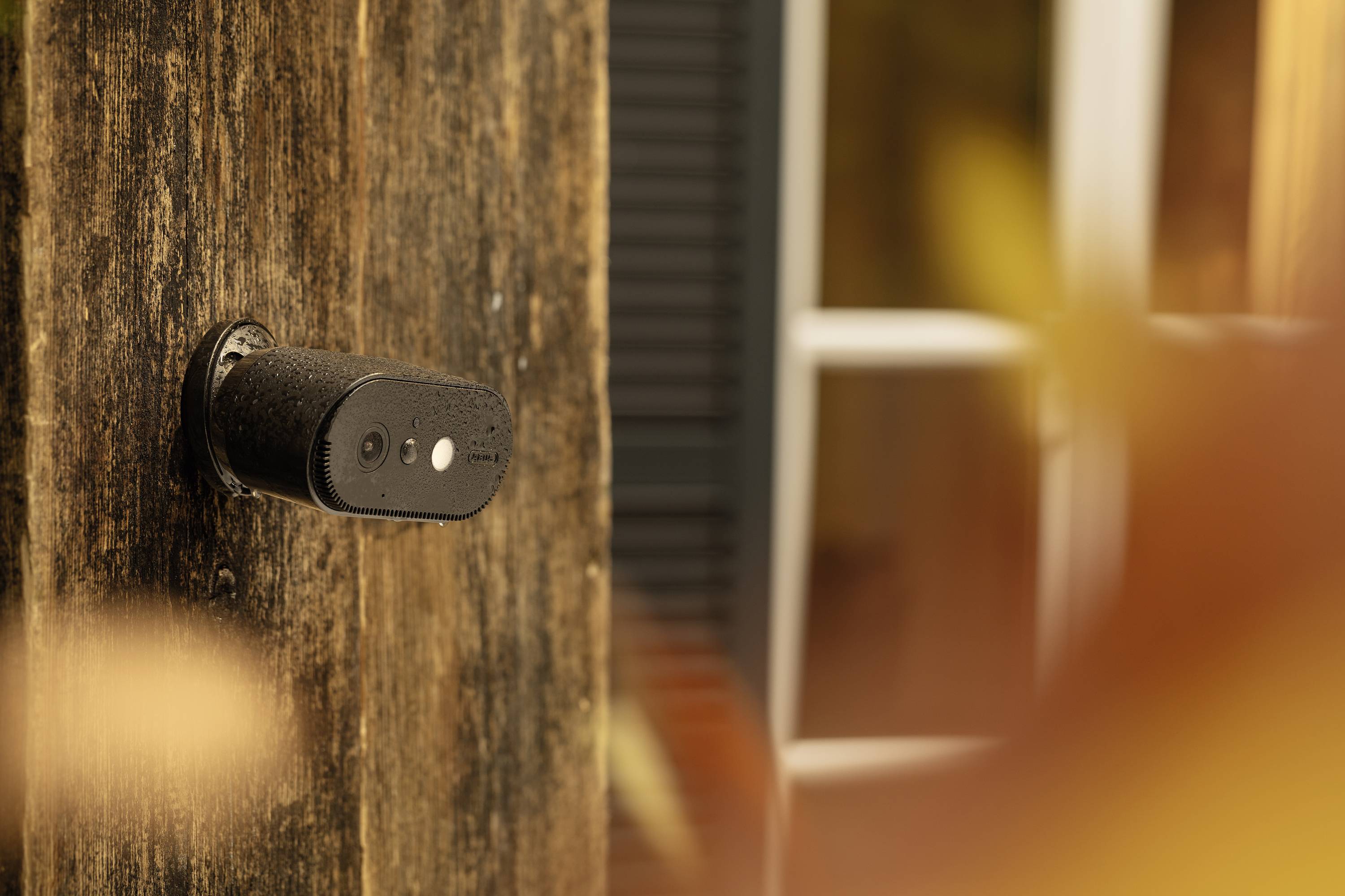 Bezpečnostní kamera je namontována z boku na dřevěné stěně, přičemž v pozadí je okno.