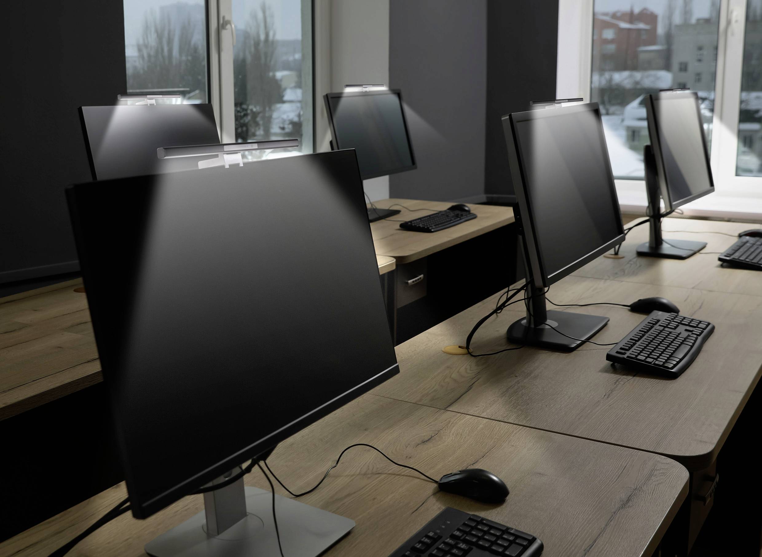 Několik prázdných počítačových pracovišť v moderní kanceláři s velkými monitory, klávesnicemi a myšmi na dřevěných stolech.