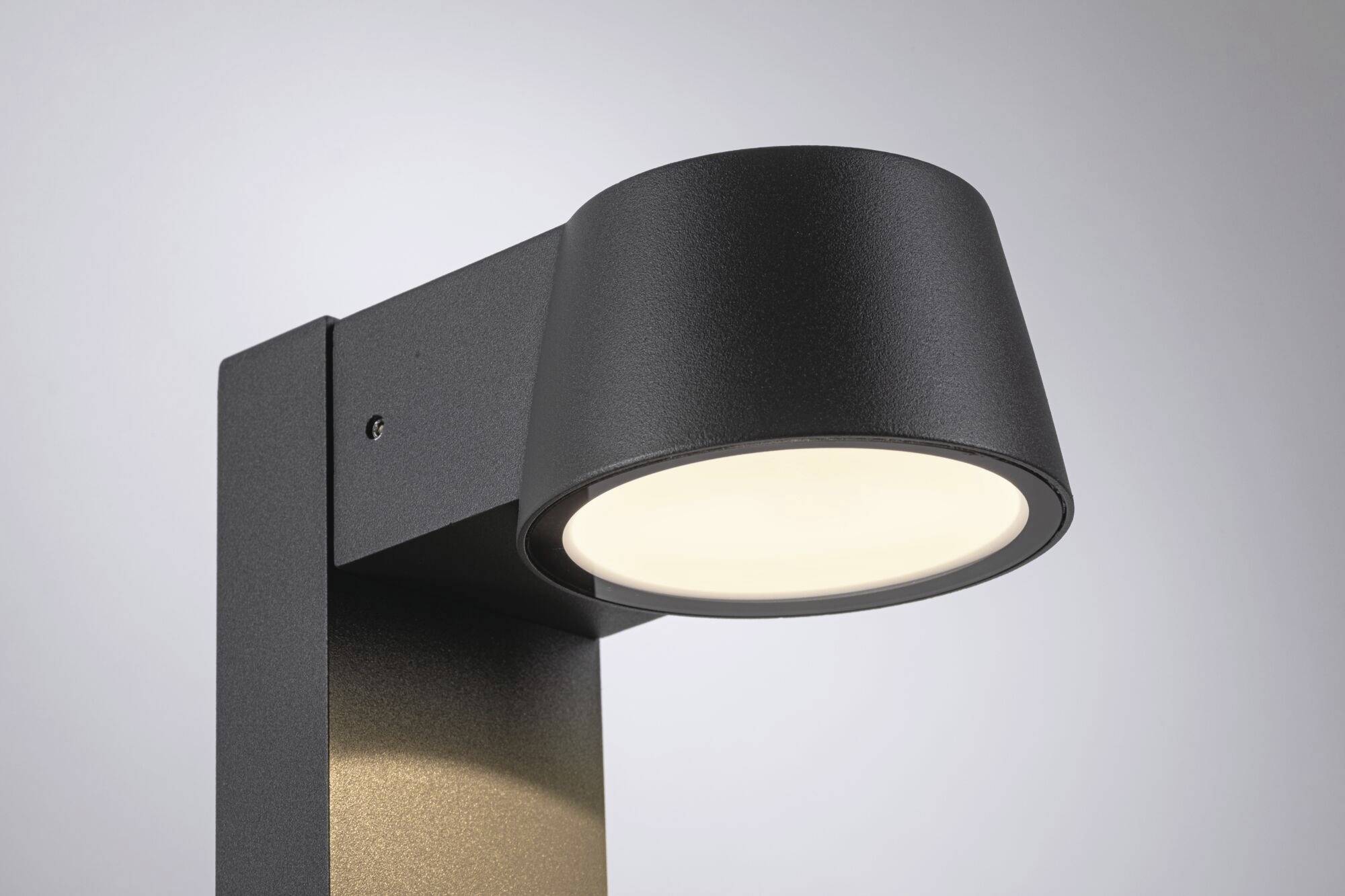 Moderní černá venkovní lampa s kulatým stínítkem vyzařuje jasné světlo. Upevněná na čtvercovém kovovém sloupu před šedým pozadím.