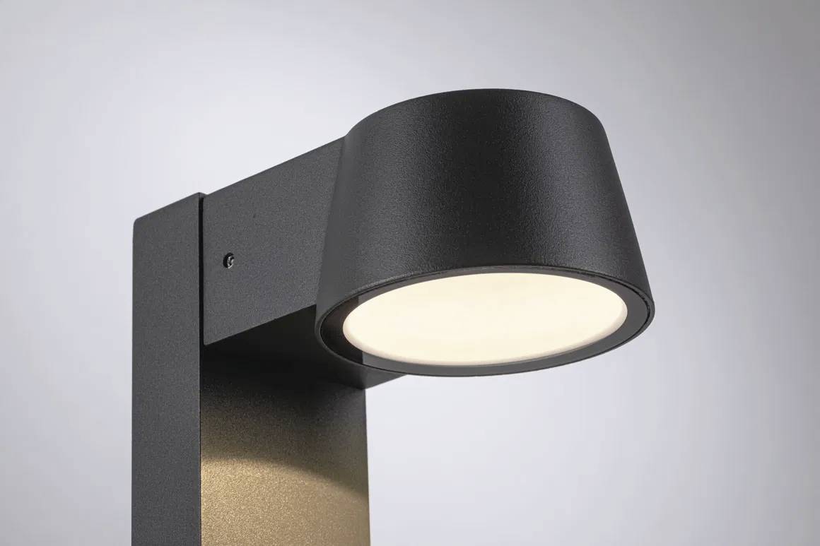 Černá, moderní venkovní lampa s kulatou, dolů orientovanou svítící hlavicí na obdélníkové konzoli, před šedým pozadím.