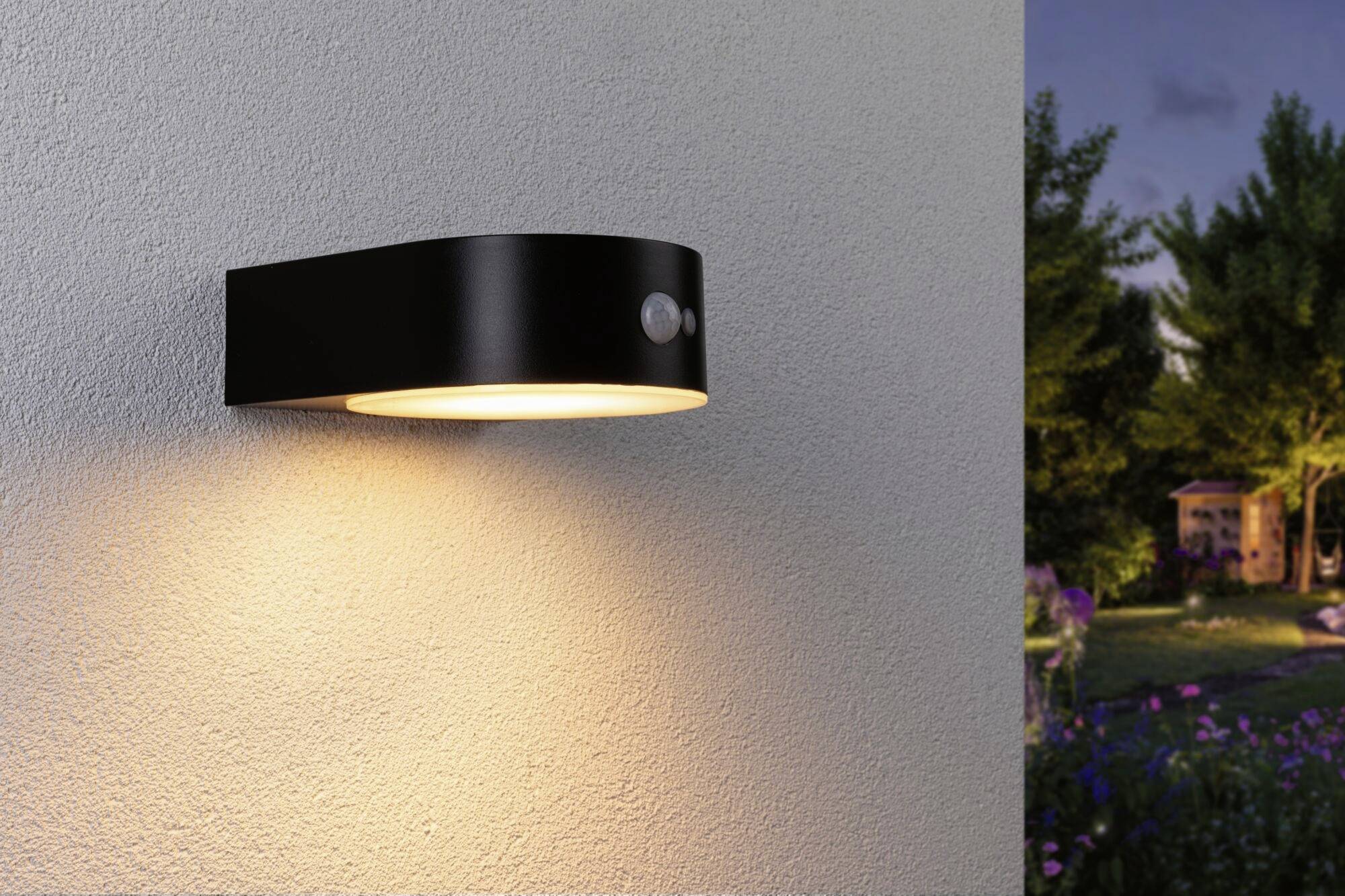Nástěnná lampa s černým pouzdrem svítí na stěně. Vpravo v pozadí zahrada se stromy a květinami za soumraku.