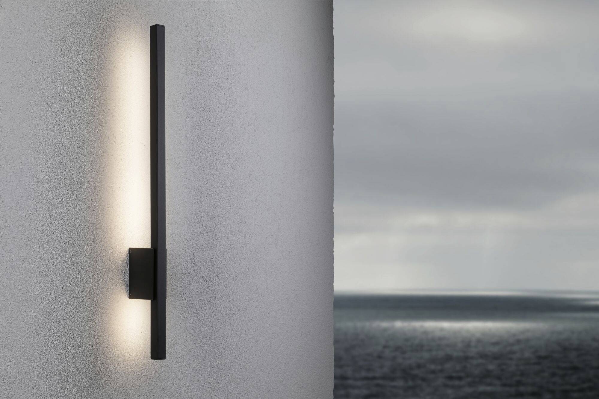 Nástěnná lampa na bílé venkovní stěně, osvětlená za zatažené oblohy s výhledem na moře v pozadí.