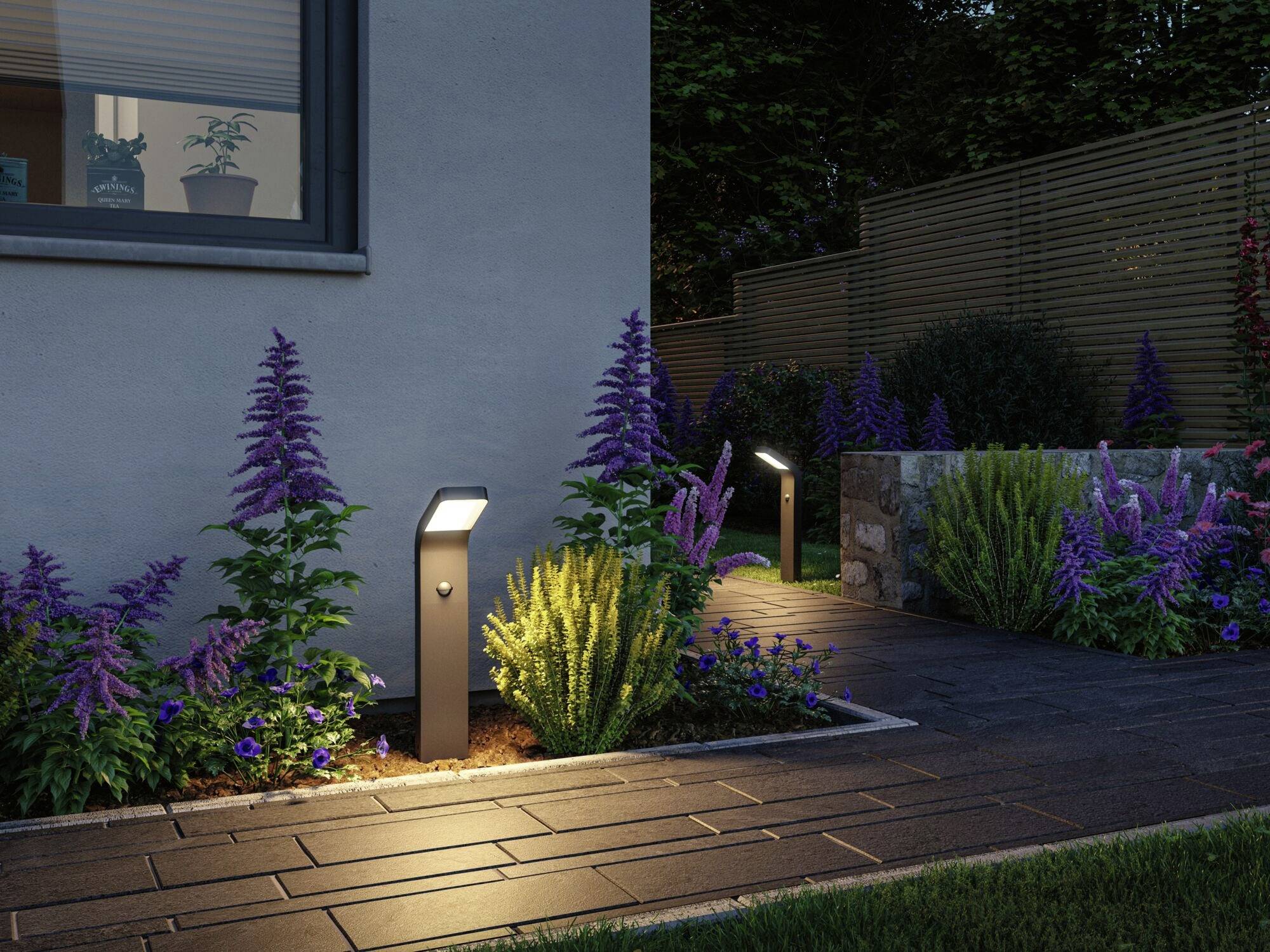 Osvětlená zahradní cesta s moderními lampami, lemovaná kvetoucími rostlinami a pečlivě udržovaným trávníkem.