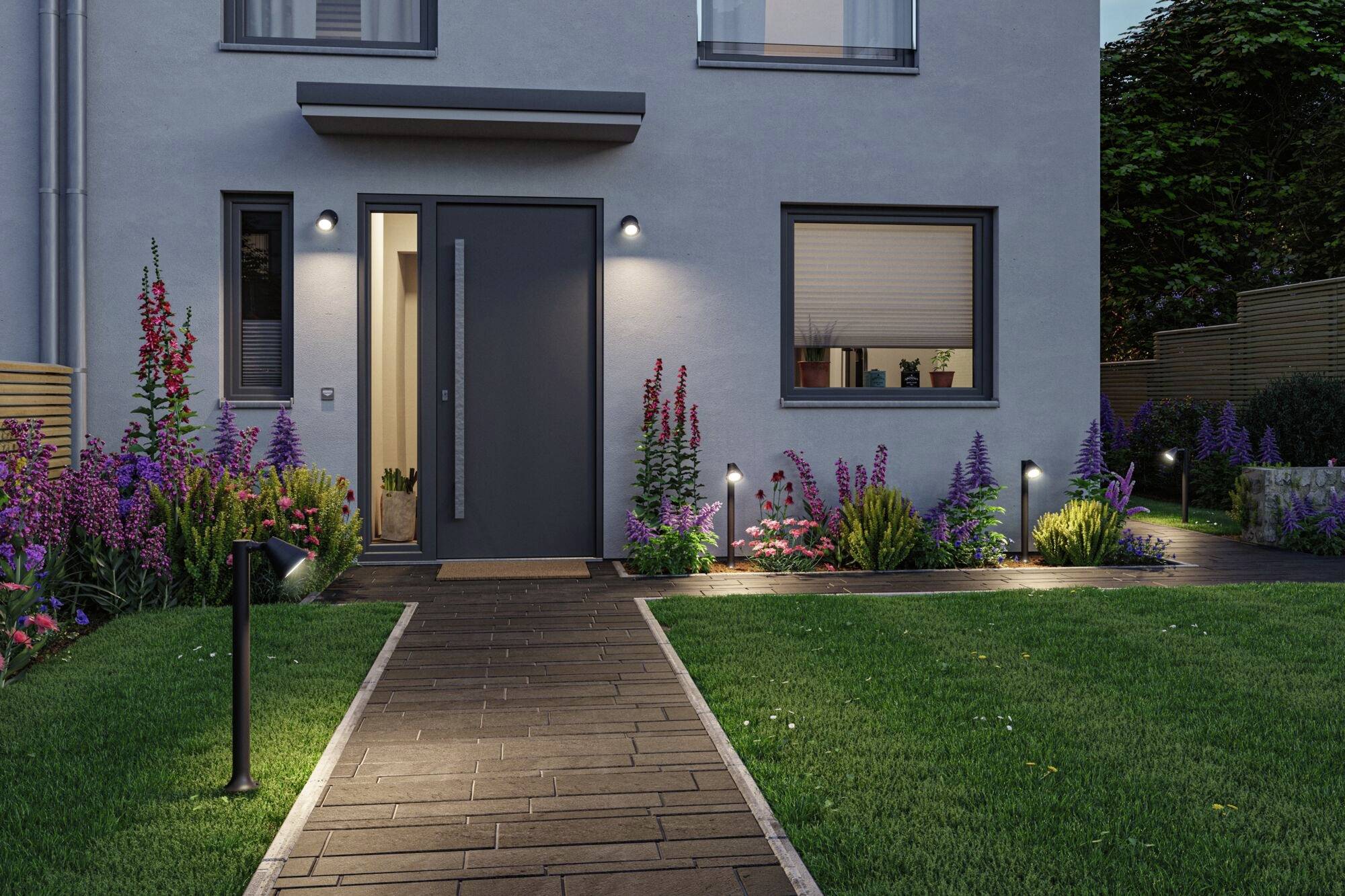 Moderní dům s osvětleným vstupem, obklopený kvetoucími rostlinami a udržovaným trávníkem.