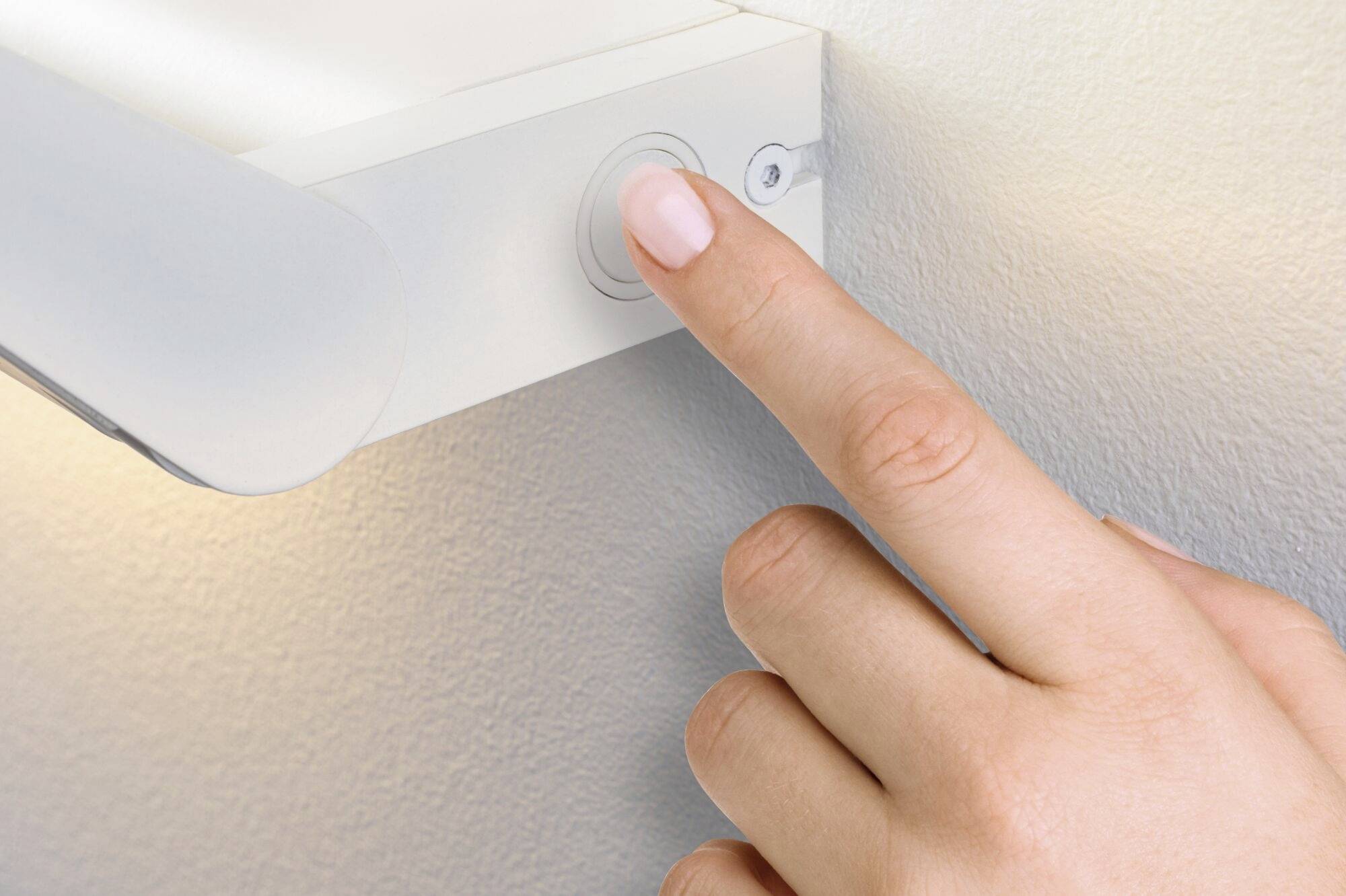 Ruka stiskává vypínač bílé nástěnné lampy, aby rozsvítila světlo. Stěna a lampa mají moderní, minimalistický design.