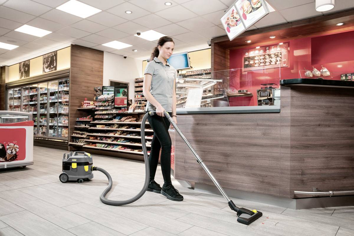 Osoba vysavující podlahu v prodejně, s policemi plnými produktů a přepážkou v pozadí, což naznačuje rutinní čištění.