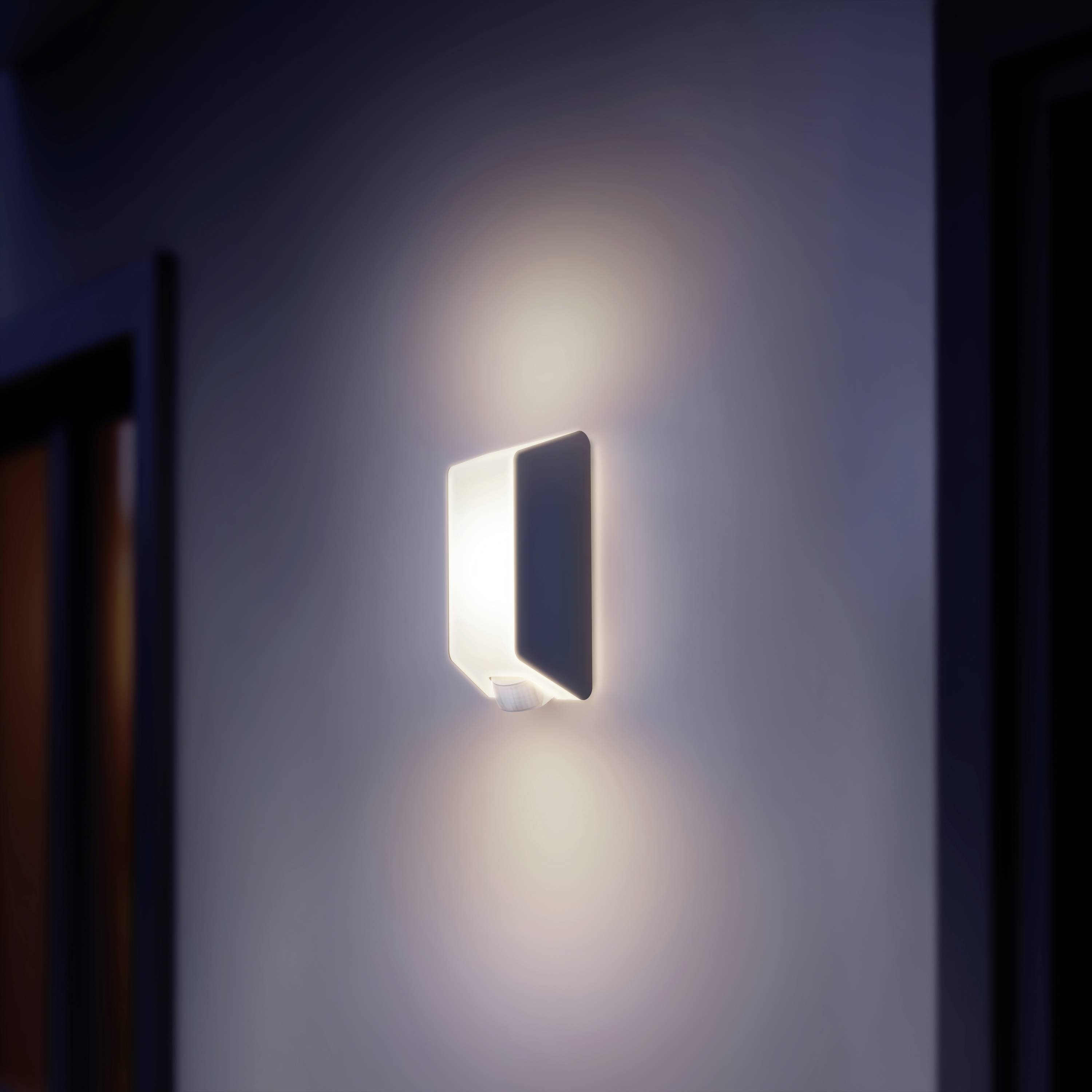 Moderní nástěnná lampa s jemným bílým světlem, upevněná na šedé zdi. Osvětluje prostor nenápadně a rovnoměrně.