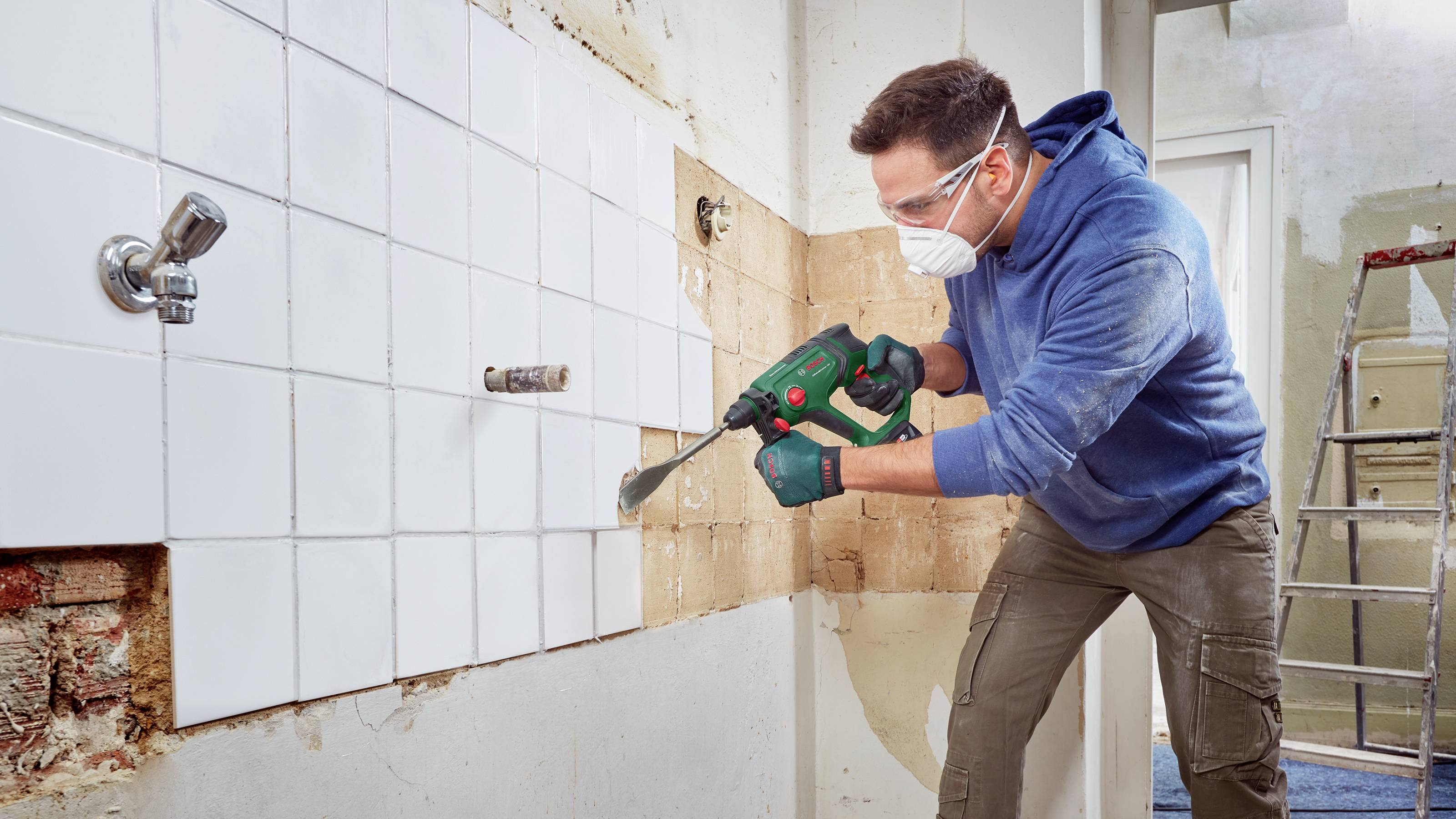 Muž odstraňuje bílé obklady ze zdi elektrickým nástrojem. Má na sobě ochranné brýle a roušku. Probíhající renovační práce.