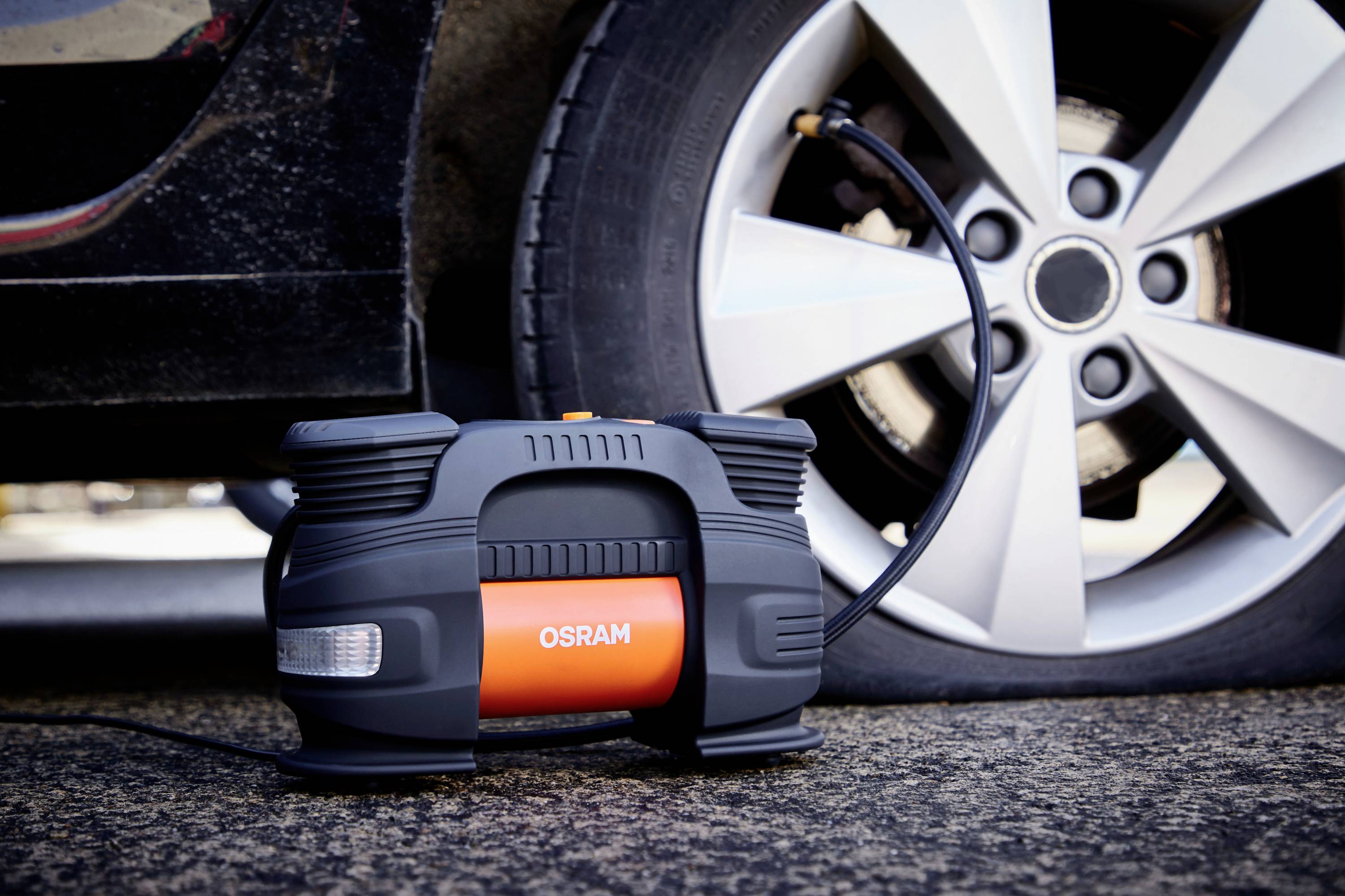 Přenosný kompresor nafukuje pneumatiku automobilu. Kompresor ukazuje značku 'OSRAM'. Pneumatika je částečně viditelná.