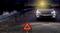 Stříbrné auto stojí v noci na dlážděné silnici se zapnutými výstražnými světly. Před ním je umístěn červený výstražný trojúhelník.