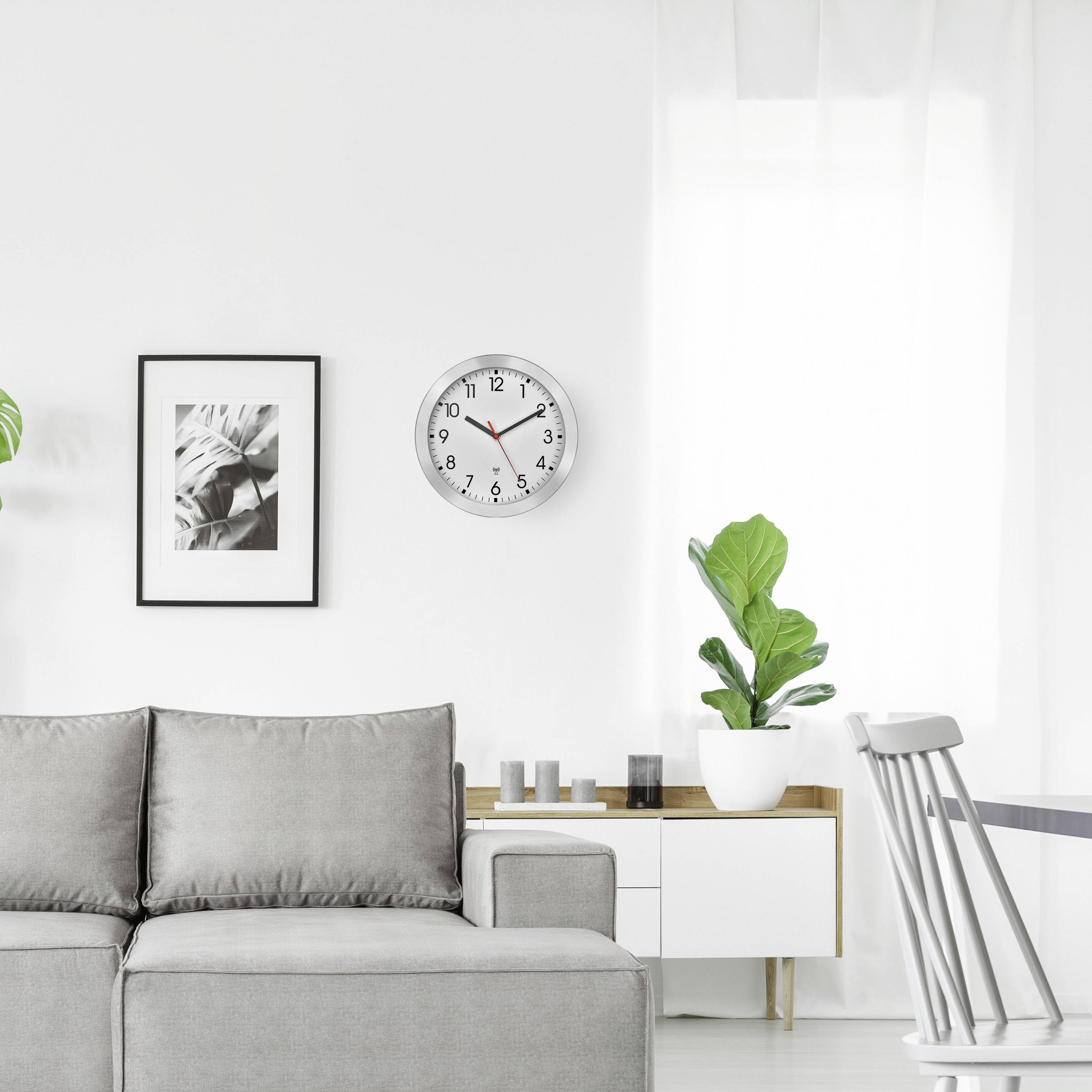 Moderní obývací pokoj s šedou pohovkou, hodiny na stěně, bílou knihovnou a pokojovými rostlinami. Světlé, minimalistické zařízení.